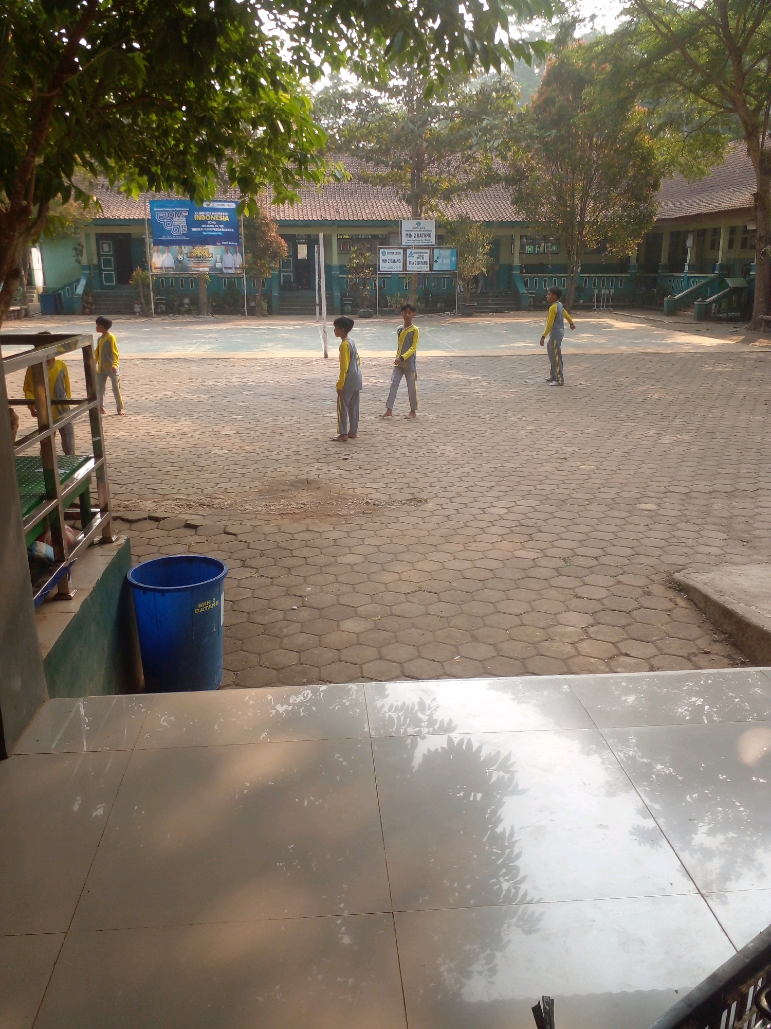 The image depicts an outdoor schoolyard with several children in yellow and gray uniforms walking on a paved surface. In the background, there is a building with a tiled roof and a banner displaying "OLIMPIADE NASTIONAL INDONESIA". A blue trash can is visible on the left side of the image. The scene appears to be in the city of Banyuputih, Indonesia, likely during the daytime.