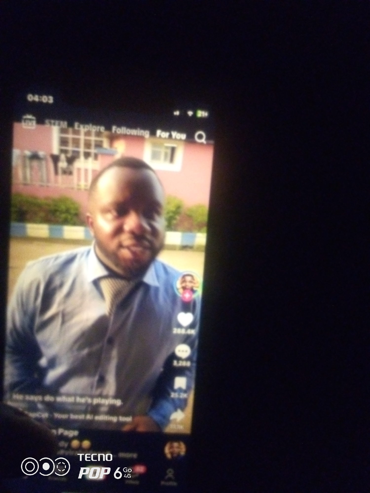 The image is a close-up shot of a smartphone screen displaying a social media application, likely TikTok, given the interface elements. The screen shows a man with dark skin and short hair, wearing a light blue collared shirt and a tie. He is looking slightly to his left. The background appears to be an outdoor scene with a pink building and some greenery.
On the screen, various app elements are visible:
- At the top left, "04:03" is displayed, likely indicating the time or video duration.
- To the right of the time, there are icons for "STEM," "Explore," "Following," and "For You."
- A search icon (magnifying glass) is present on the top right.
- To the right of the man's image, there are several icons with numbers indicating engagement metrics: a heart icon with "288.4K," a comment icon with "1,288," and a share icon with "25.2K."
- Below the man, there is text that reads "He says do what he's playing." and "Your best AI editing tool."
- The bottom of the screen displays branding for "TECNO POP 6 Go 4G" along with several circular graphic elements.
There is no information in the image to suggest the location is Howayi, Nigeria. The provided description is based solely on the visual content of the image.