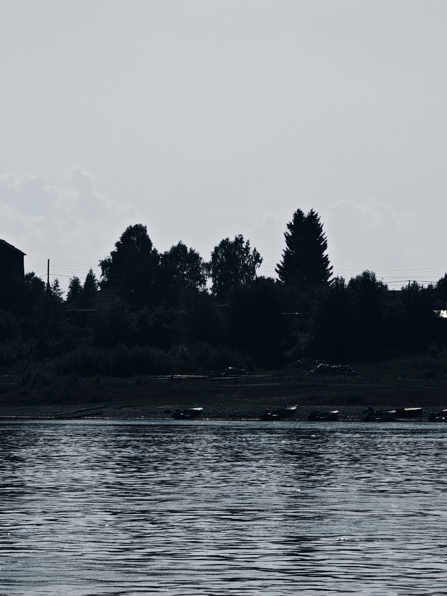 The image captures a serene view of the river in Verkhnepashino, Russia. In the foreground, the water is rippled, reflecting the overcast sky. The middle ground shows a densely wooded shoreline, silhouetted against the light. Various trees of different shapes and sizes form a dark, undulating line. Among the trees, hints of structures can be discerned, including a building on the far left and what appears to be a roof with corrugated siding further in. Power lines are visible stretching across the scene, supported by poles. On the bank, several small boats are pulled ashore, their shapes dark and indistinct. The overall atmosphere is tranquil and slightly melancholic, with the muted tones and the silhouettes creating a sense of quietude.