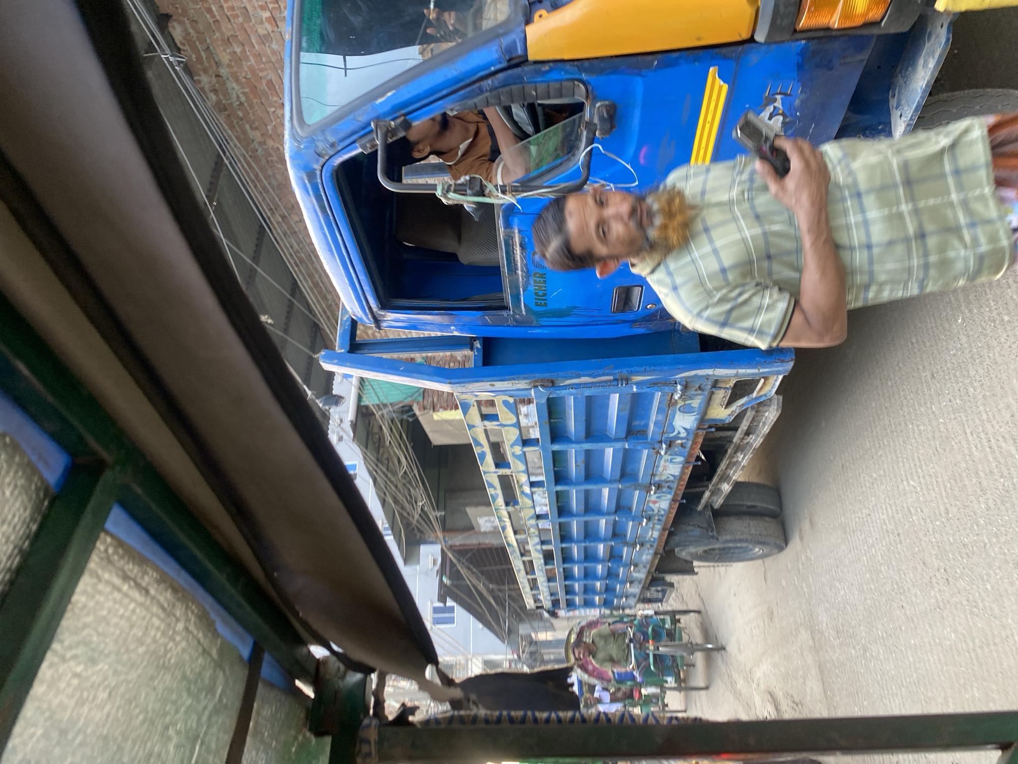 The image depicts a street scene in Dhaka, Bangladesh, with a blue Eicher truck parked on the roadside. A man in a plaid shirt is standing next to the truck, holding a mobile phone. The driver of the truck is inside the cabin. In the background, there are other vehicles, including cycle rickshaws, and buildings lining the street. The scene is viewed from a low angle, possibly from inside another vehicle.