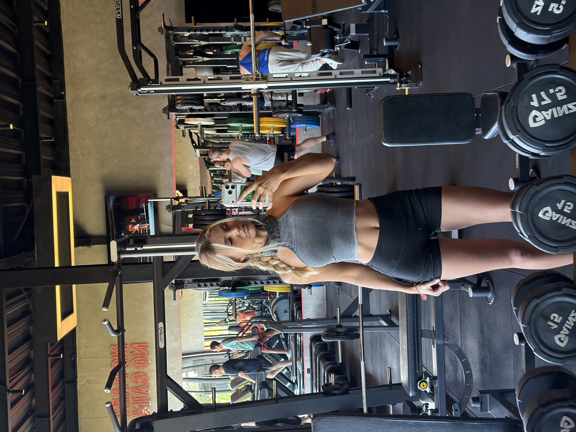 The image depicts a woman taking a mirror selfie in a gym located in North Kuta, Indonesia. The woman is wearing a grey crop top and black shorts. She has blonde hair styled in a braid. In the background, other gym-goers can be seen using exercise equipment, including treadmills and weight machines. The gym features industrial-style lighting and concrete walls. The overall scene is that of a modern fitness facility.