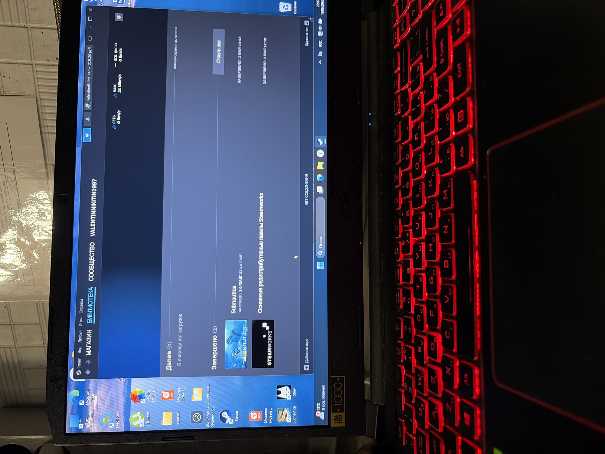 The image shows a close-up of a laptop screen displaying the Steam game client. The Steam interface is visible, with the user's library categorized into "Further (0)" and "Completed (2)". Under "Completed", two items are listed: "Subnautica" and "Main redistributable packages Steamworks". The top of the screen shows the username "valentininitkin1997" and a currency amount of "219,29 руб". The laptop's keyboard, illuminated with red backlighting, is in the foreground. The overall environment appears to be indoors, with a textured wall in the background. No specific location in a city is identifiable.