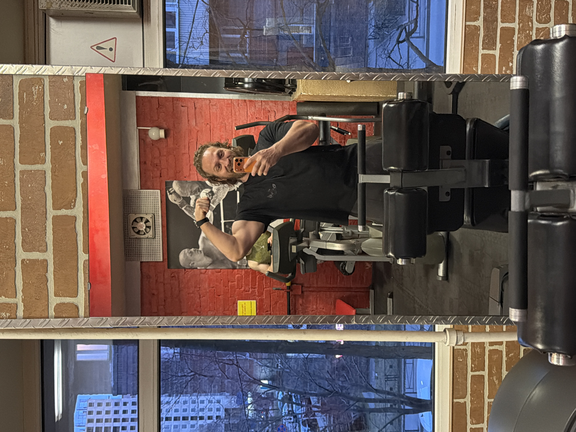 A man is taking a selfie in a gym. He is holding an orange smartphone and flexing his bicep. The gym has red brick walls and a mirror. The reflection in the mirror shows a window with a view of the city of Saint Petersburg, Russia.