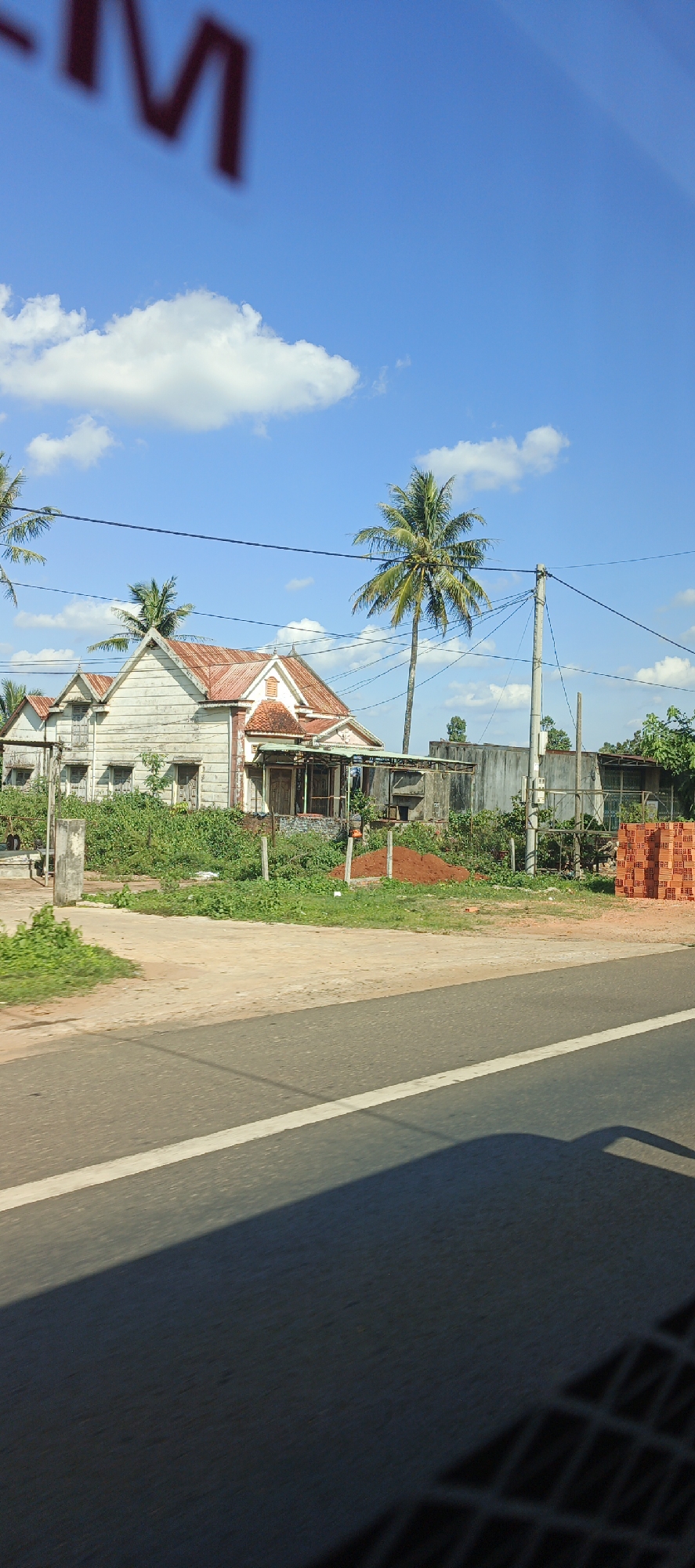 This is a vertical, eye-level shot of a rural scene in Chư Sê, Vietnam, under a bright blue sky with scattered white clouds. The foreground features a paved road with a white line and a dark shadow cast from a vehicle. Beyond the road, there is a slightly overgrown area with green vegetation and a dirt path. A group of buildings is situated in the midground. The main building is a weathered, light-colored wooden house with a reddish-brown metal roof and arched window frames. To the right of this house are several smaller structures, including one with a metal roof and what appears to be a concrete wall. Piled high to the far right are stacks of orange bricks. Several tall palm trees dot the landscape, with one prominent palm tree in the center of the image. Electrical wires crisscross the scene above the buildings and trees. In the blurred background at the top of the image, red lettering is visible against the sky. The overall impression is one of a quiet, slightly dilapidated rural settlement.