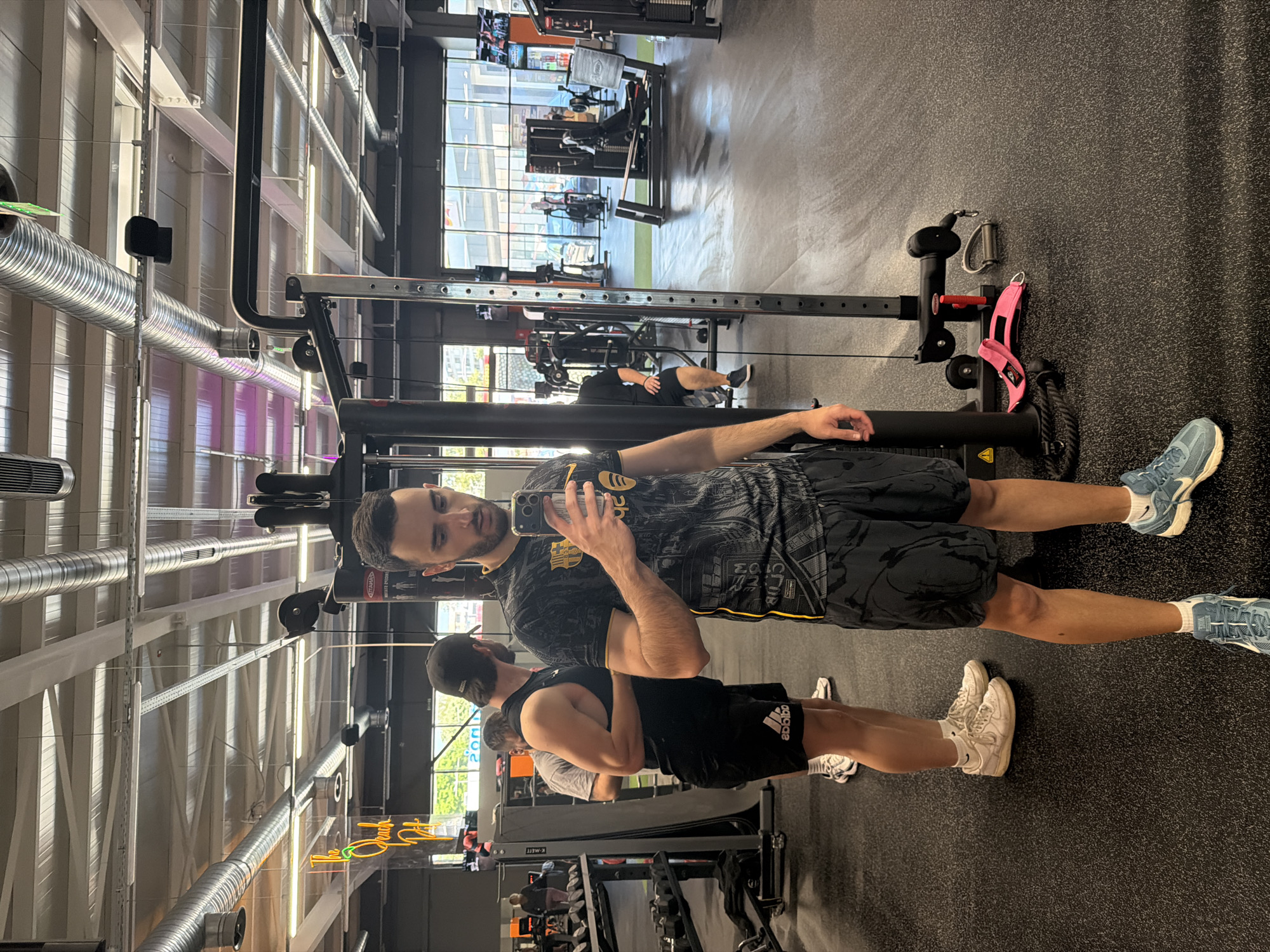 The image shows the interior of a gym. Two males are visible, one taking a selfie. The gym contains various exercise machines, including a cable machine in the foreground and other weight machines in the background. The location appears to be the PULSE FITNESS & SPA Plovdiv, Bulgaria, identified by the green and orange sign reflected in the phone screen. The floor is dark gray, and the ceiling features exposed ductwork and lighting fixtures. Outside the windows, indistinct buildings and greenery can be seen.