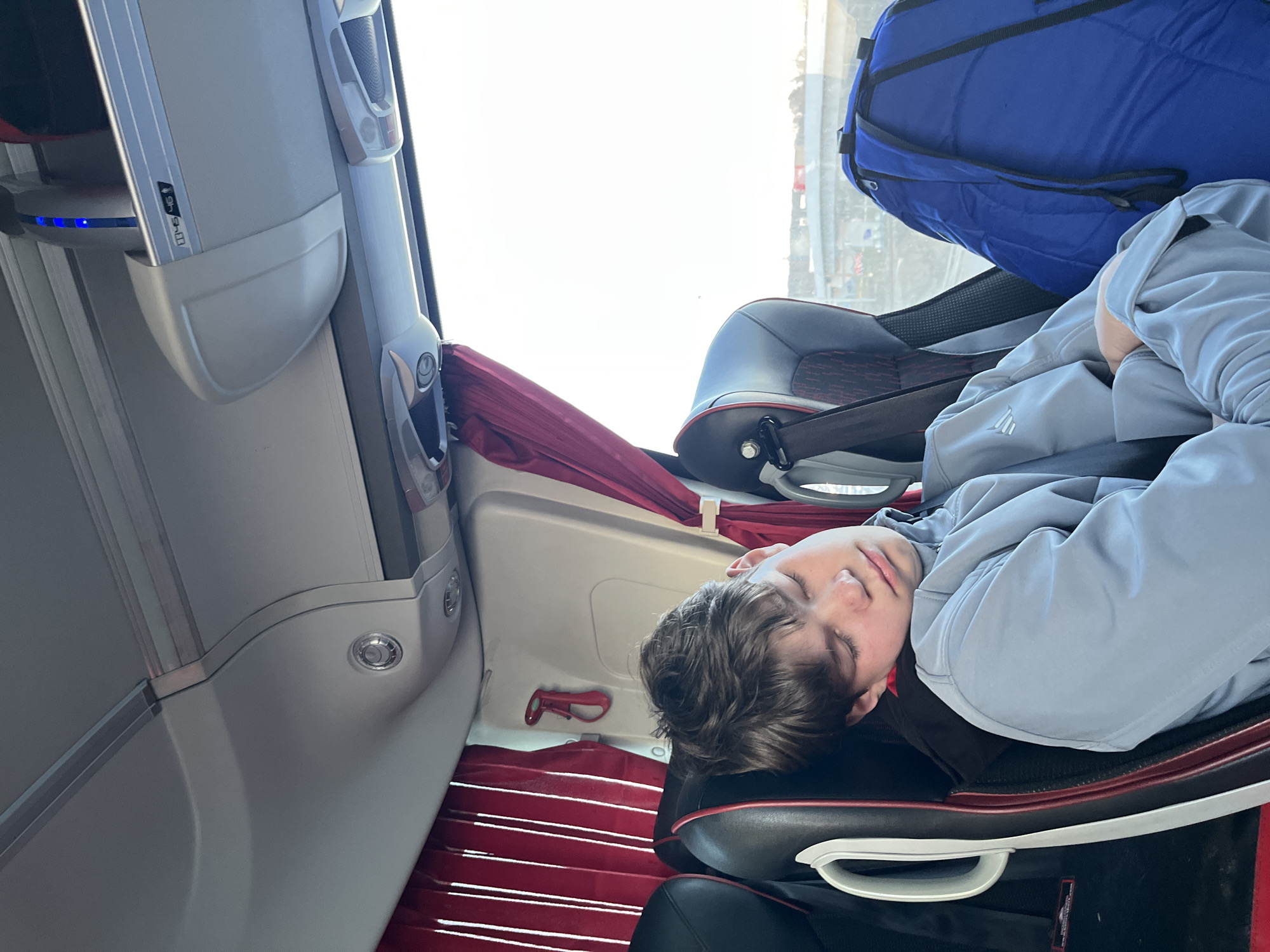 A young person is sleeping in a seat on a bus. The bus has luggage storage above the seats, and a large blue duffel bag is visible above the sleeping person. The seat is black with red trim. The bus interior is mostly white with red accents. The bus appears to be in motion. The city is not evident from this image.