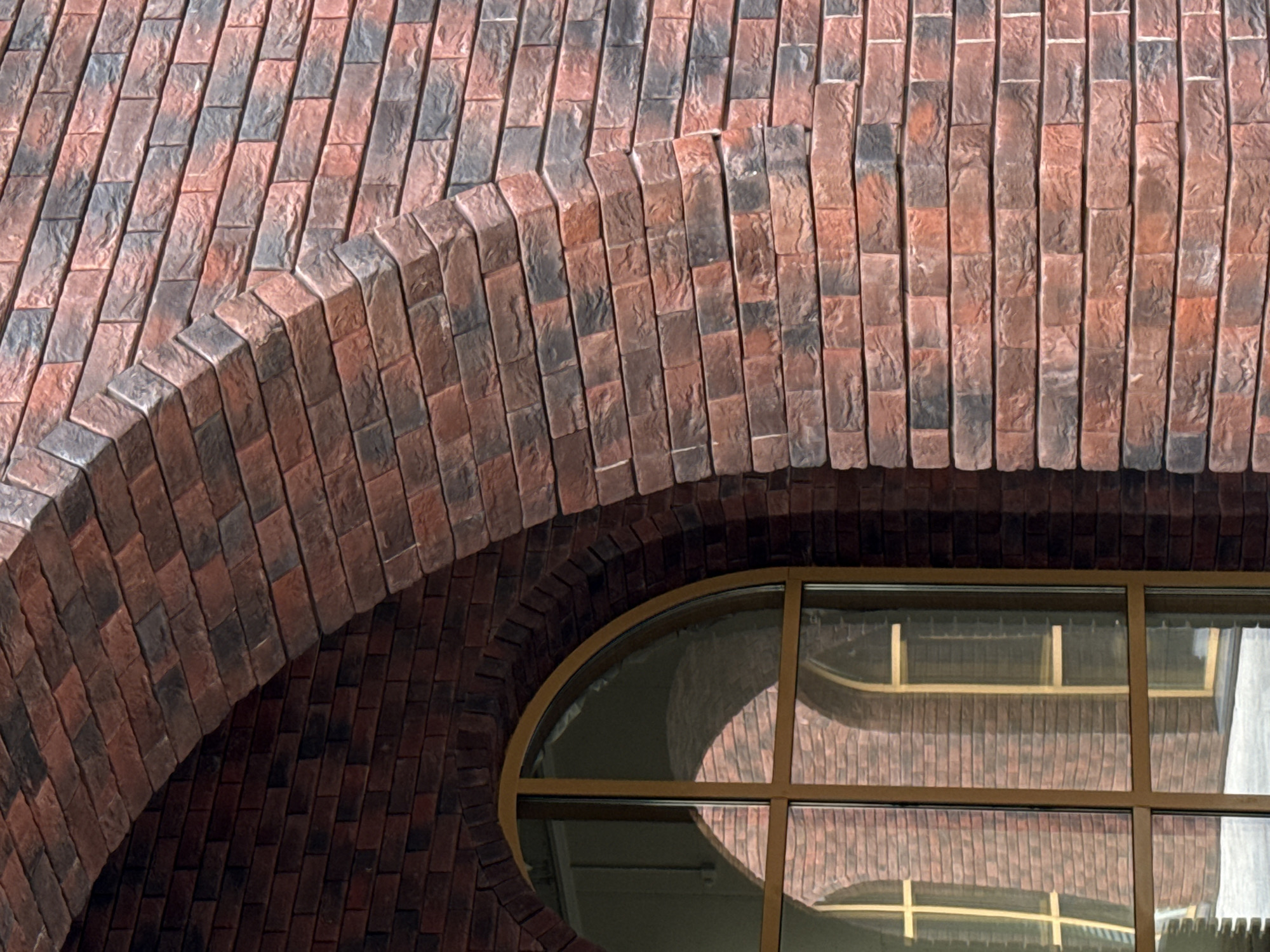 Close-up view of the exterior facade of the National Library of the Udmurt Republic in Izhevsk, Russia. The facade features red-brown brickwork arranged in both straight and curved sections, forming arches. A round window with a golden frame is visible, reflecting the brickwork of the surrounding building.