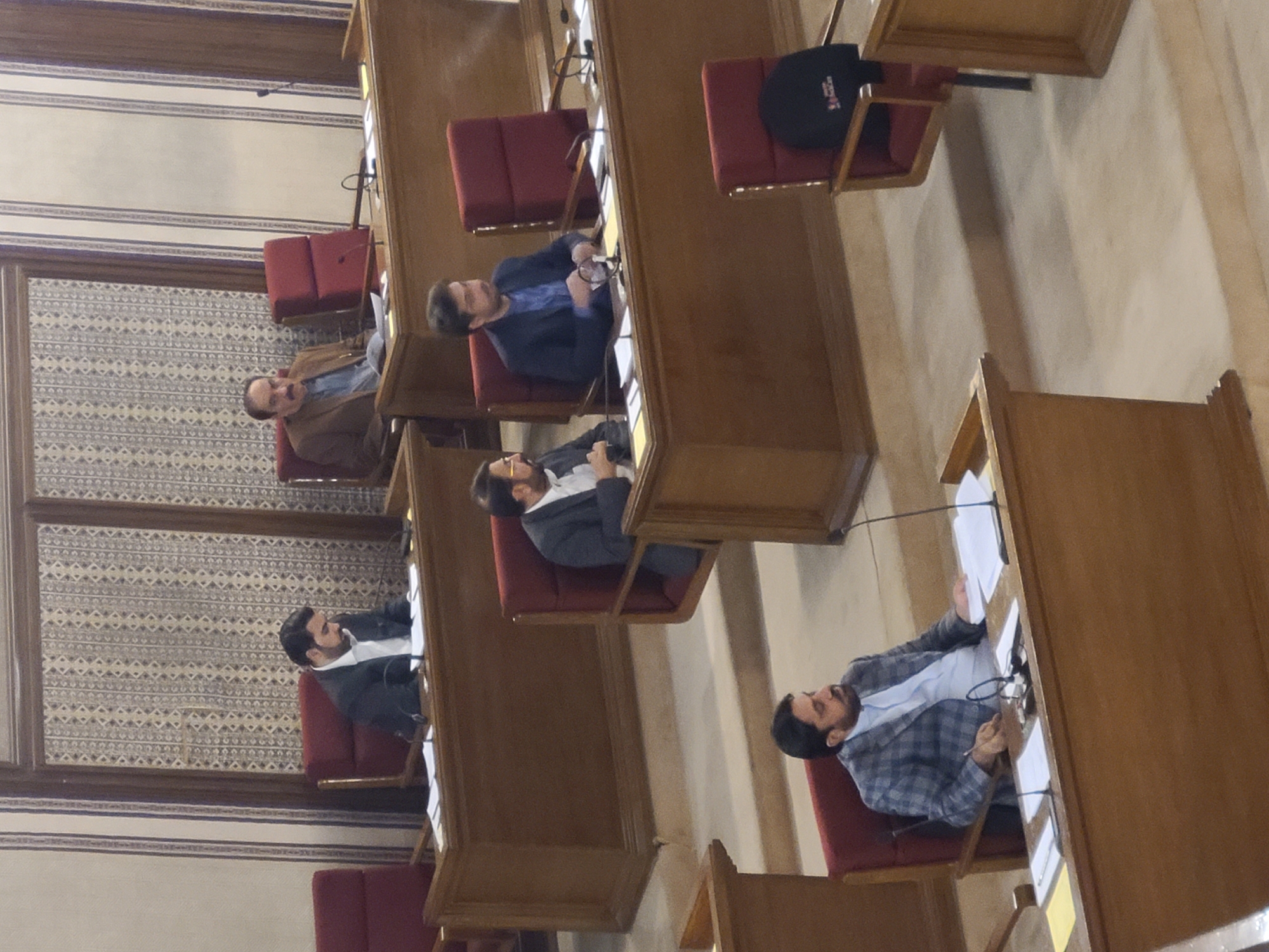 The image shows four men seated in what appears to be a courtroom or legislative chamber. The men are wearing suits and are seated at wooden desks with microphones. The location is indicated as Quetta City Tehsil, Pakistan. The environment consists of paneled walls and patterned wallpaper. The men appear to be engaged in a formal meeting or session. Some have papers in front of them, suggesting they are reviewing documents. One man is actively taking notes or writing.