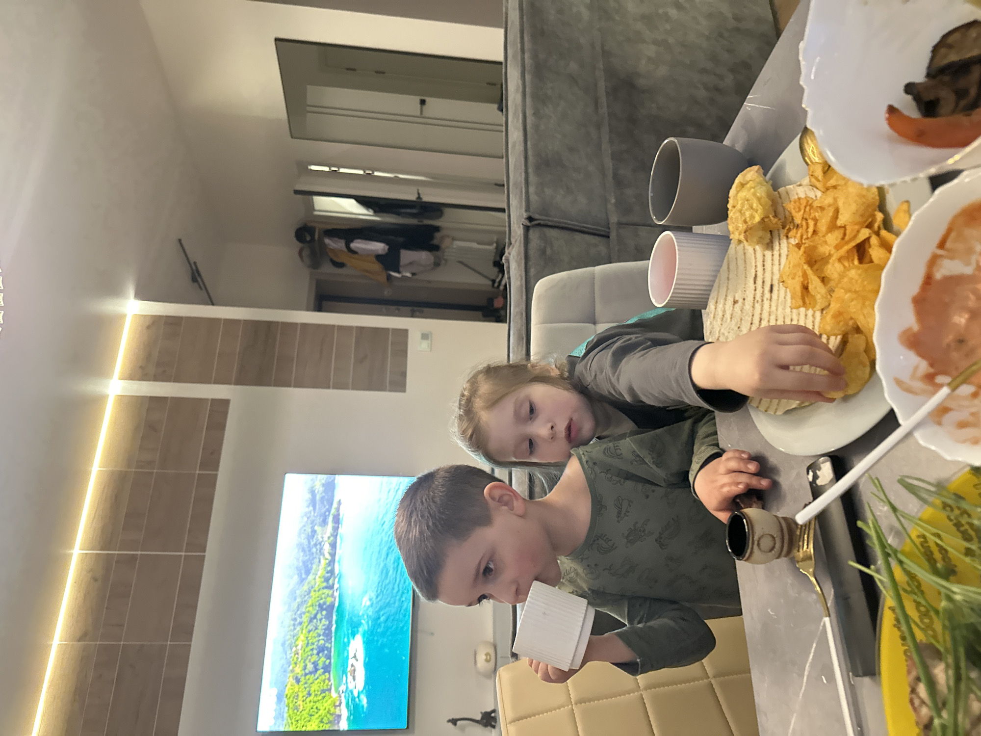 The image shows two children at a table with food. The younger child, a girl with light hair, is reaching for potato chips. The older child, a boy, is holding a white container. Food items on the table include chips, tortillas, and what appears to be a sauce. The location appears to be indoors, with a large TV displaying a beach scene, suggesting a residential setting. There is no immediate evidence to confirm the city as Chaikovsky, Russia.