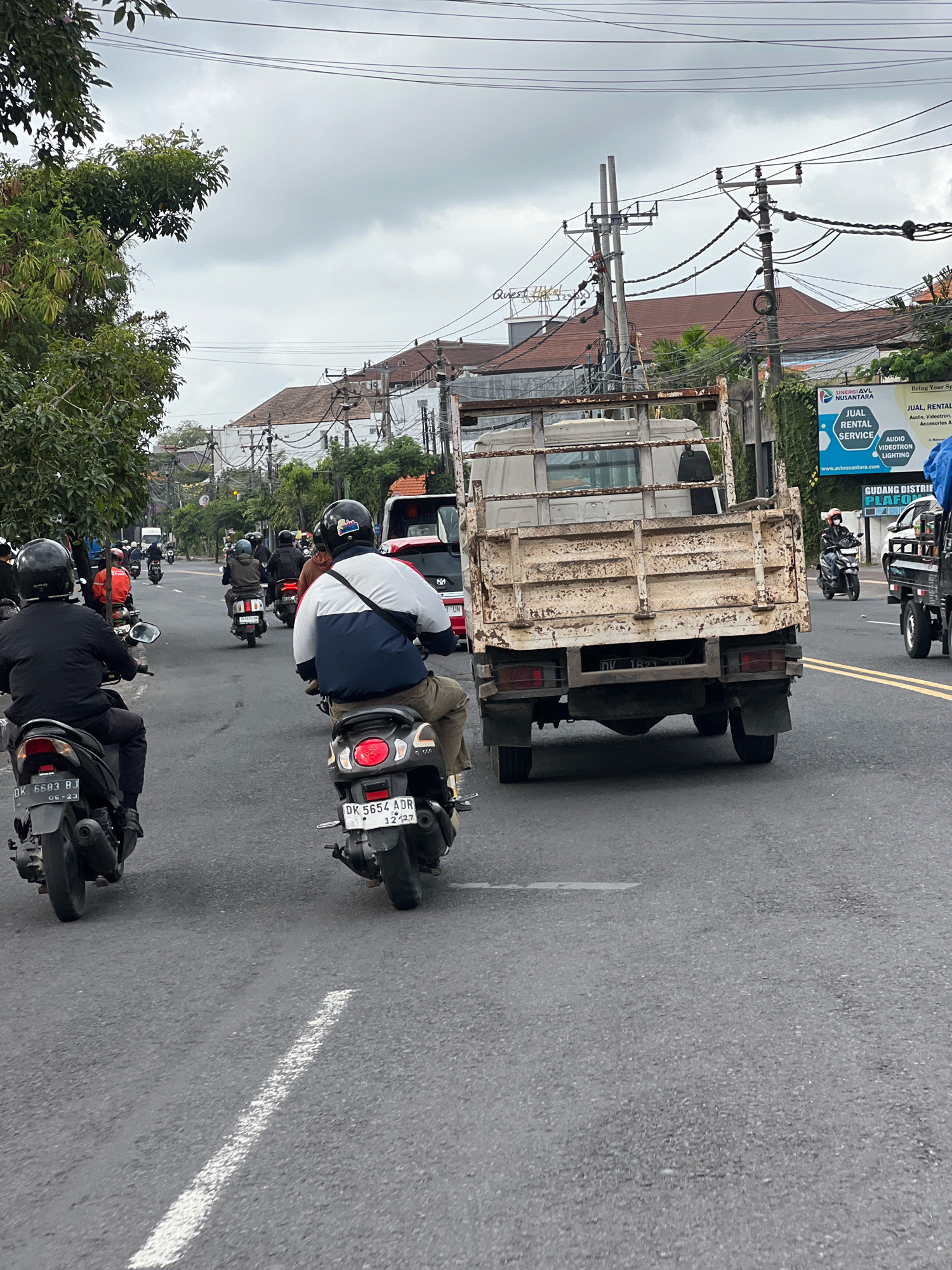 The image depicts a street scene in Denpasar, Indonesia, likely on a cloudy day. The foreground shows the rear view of two motorcycles. The motorcycle on the left has a black seat, black handlebars, and a license plate that reads "DK 6683 BJ 06-23". The rider is wearing a black helmet and a black jacket. The motorcycle on the right has a darker color, a license plate that reads "DK 5654 ADR 12.27", and its taillight is illuminated red. The rider is wearing a helmet and a jacket with white, navy, and red sections.
In the middle ground, a rusty, light-colored dump truck is moving forward. Behind the truck, several other motorcycles and a red truck are visible, all part of the traffic flow. To the right of the truck, a blue and white billboard advertises "JUAL RENTAL SERVICE" (for Sale Rental Service) for "AUDIO VIDEOTRON LIGHTING". Overhead, numerous power lines crisscross the sky, supported by utility poles. The street is paved and appears to be a busy urban thoroughfare. The background shows buildings with tiled roofs and lush green trees lining the street.