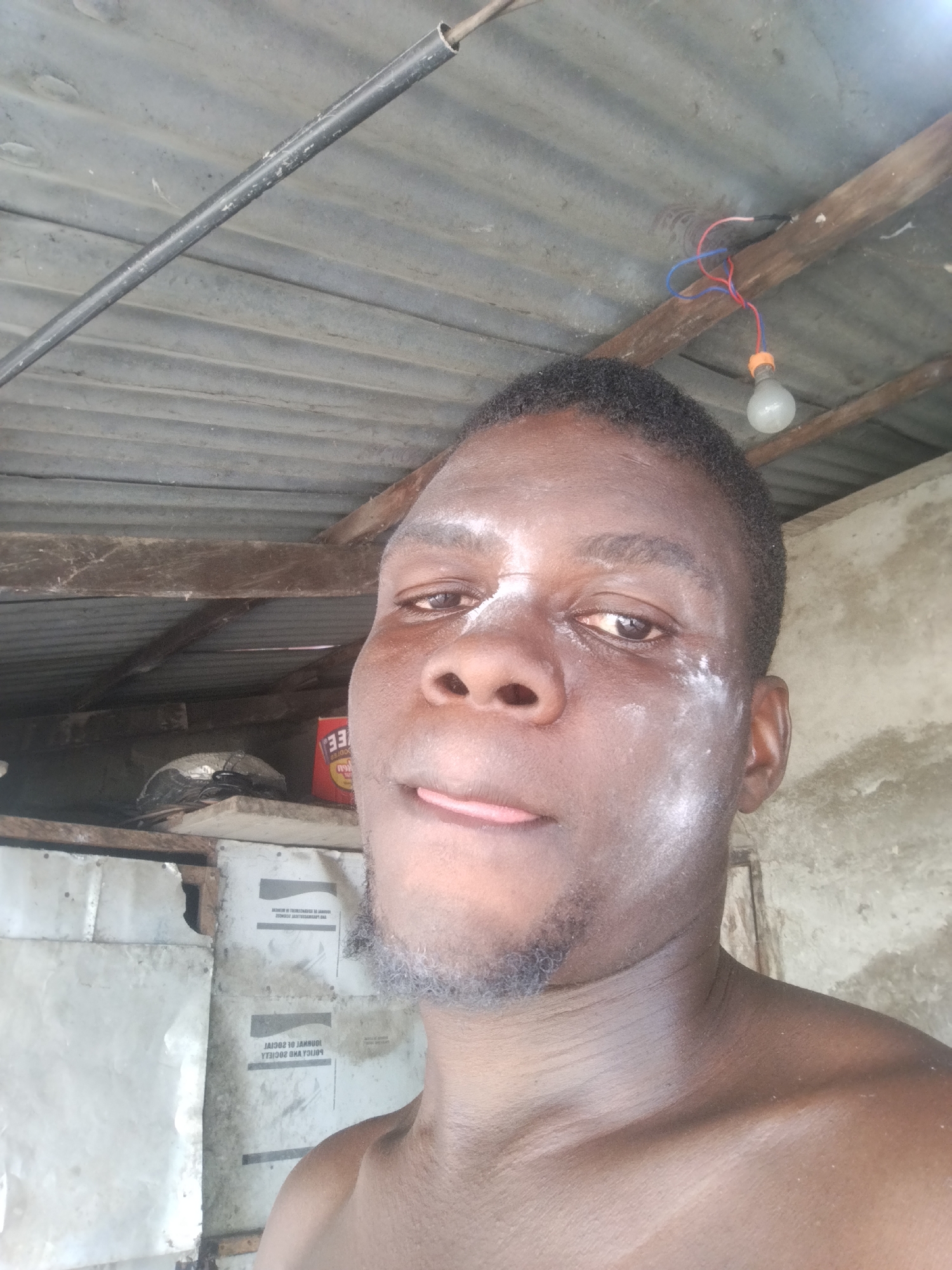 A shirtless, dark-skinned male with a goatee is centered in the frame. His face has a light dusting of what appears to be white powder. He has dark hair and is sticking his tongue out slightly.
The background includes a corrugated metal roof with a light bulb suspended from exposed wiring. There's a wooden support beam running horizontally. Boxes and various items are visible on shelves in the background. A plain, light-colored wall is seen on the right.
The location of the photograph is inside a dwelling likely located in Uyo, Nigeria, judging by the subject's ethnicity and surroundings.