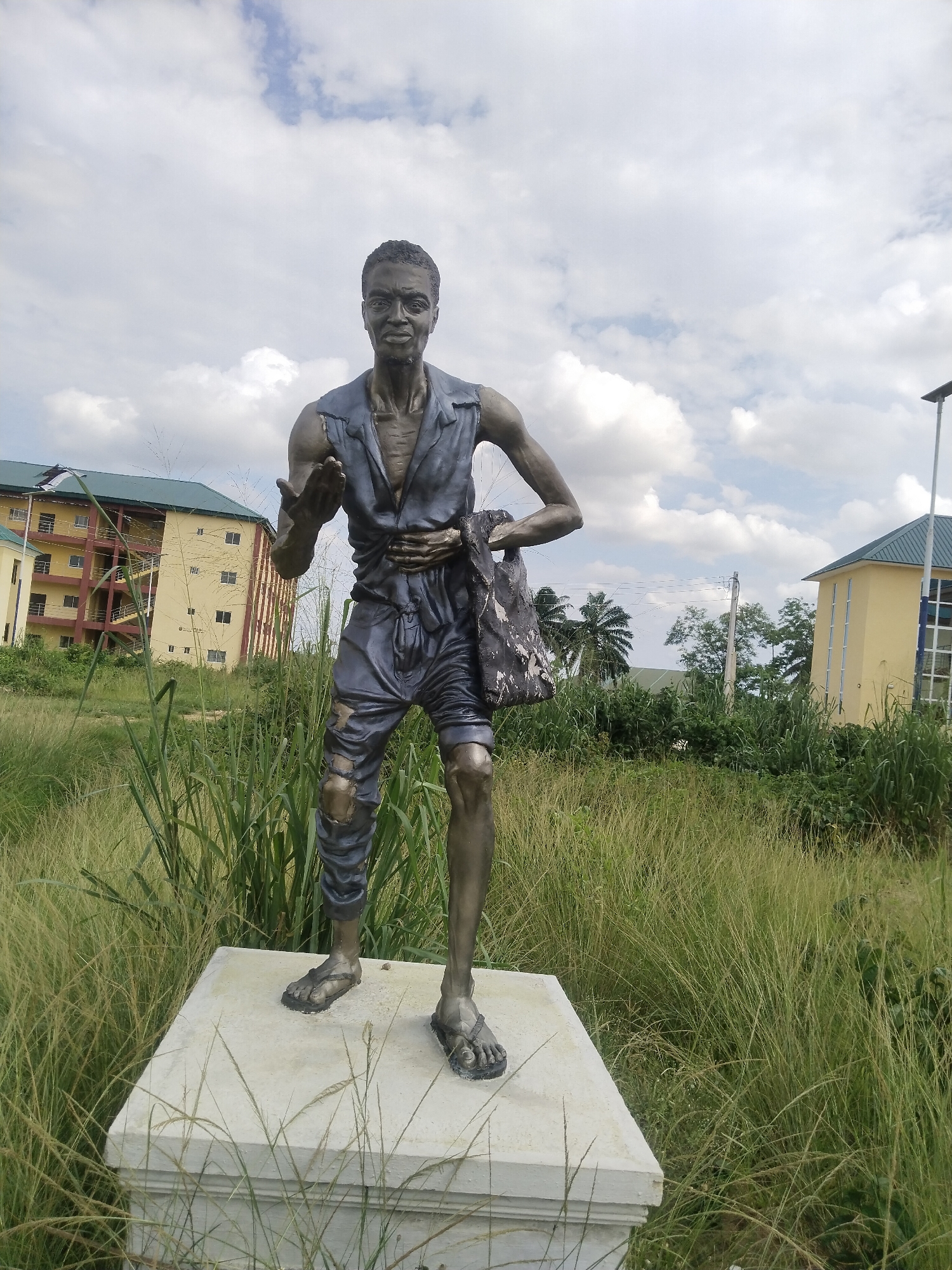 A statue of a thin, dark-skinned man stands on a concrete pedestal amidst tall grasses in Uyo, Nigeria. He wears tattered clothing: a sleeveless shirt, shorts with rips, and sandals. He is posed with one hand extended outward, as if gesturing, and a bag slung over his other shoulder. Behind the statue are two buildings, one with four stories and the other with one story, set against a backdrop of trees and a cloudy sky. The location is a grassy area in Uyo, Nigeria.