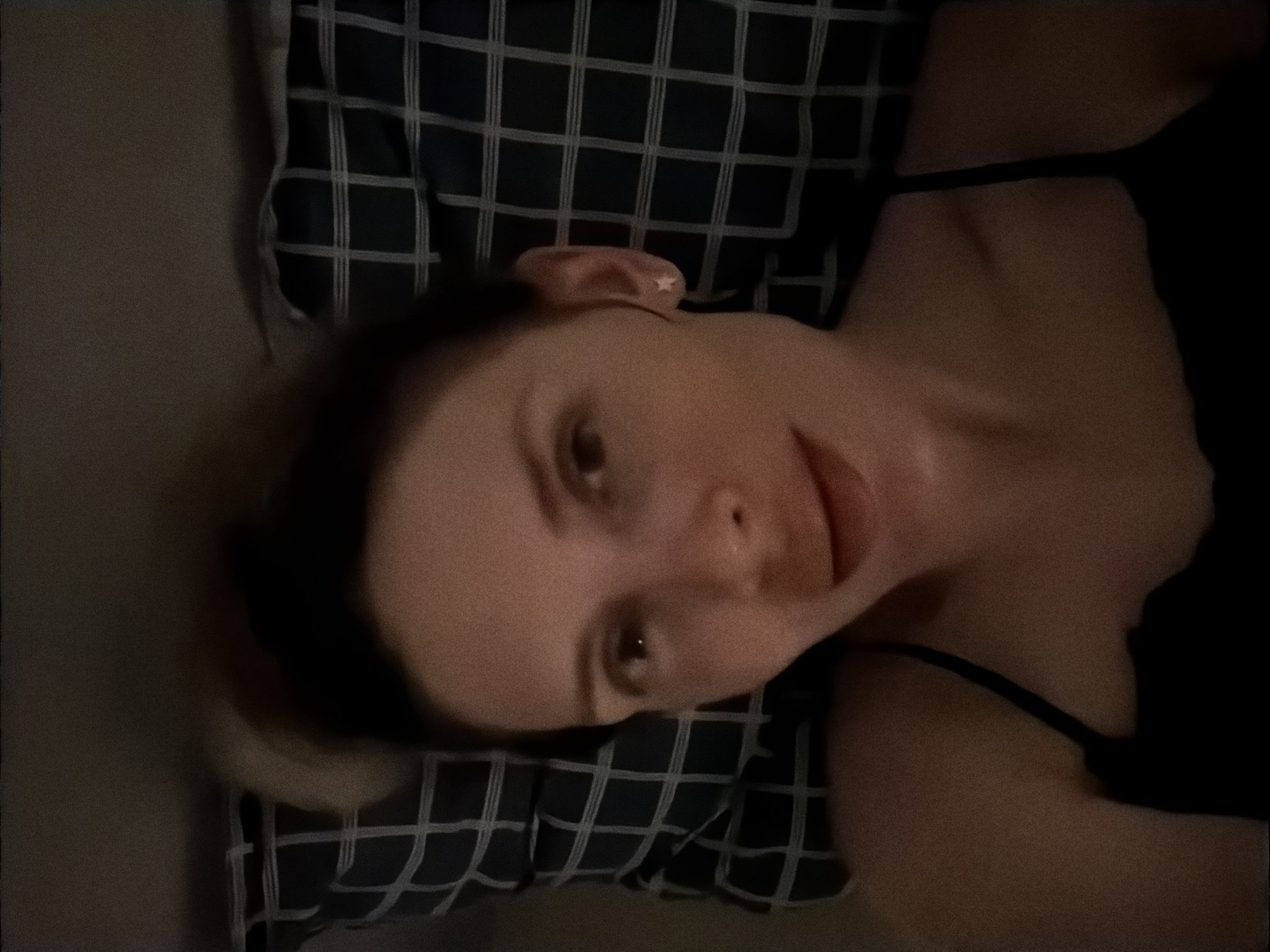 A person with pale skin and brown hair in a low ponytail is lying on a pillow with a dark blue and white grid pattern. The person is wearing a black tank top with thin straps. The image appears to be taken indoors, possibly at night. The location is not discernible from the image. There is no way to determine if the image was taken in Budva, Montenegro.