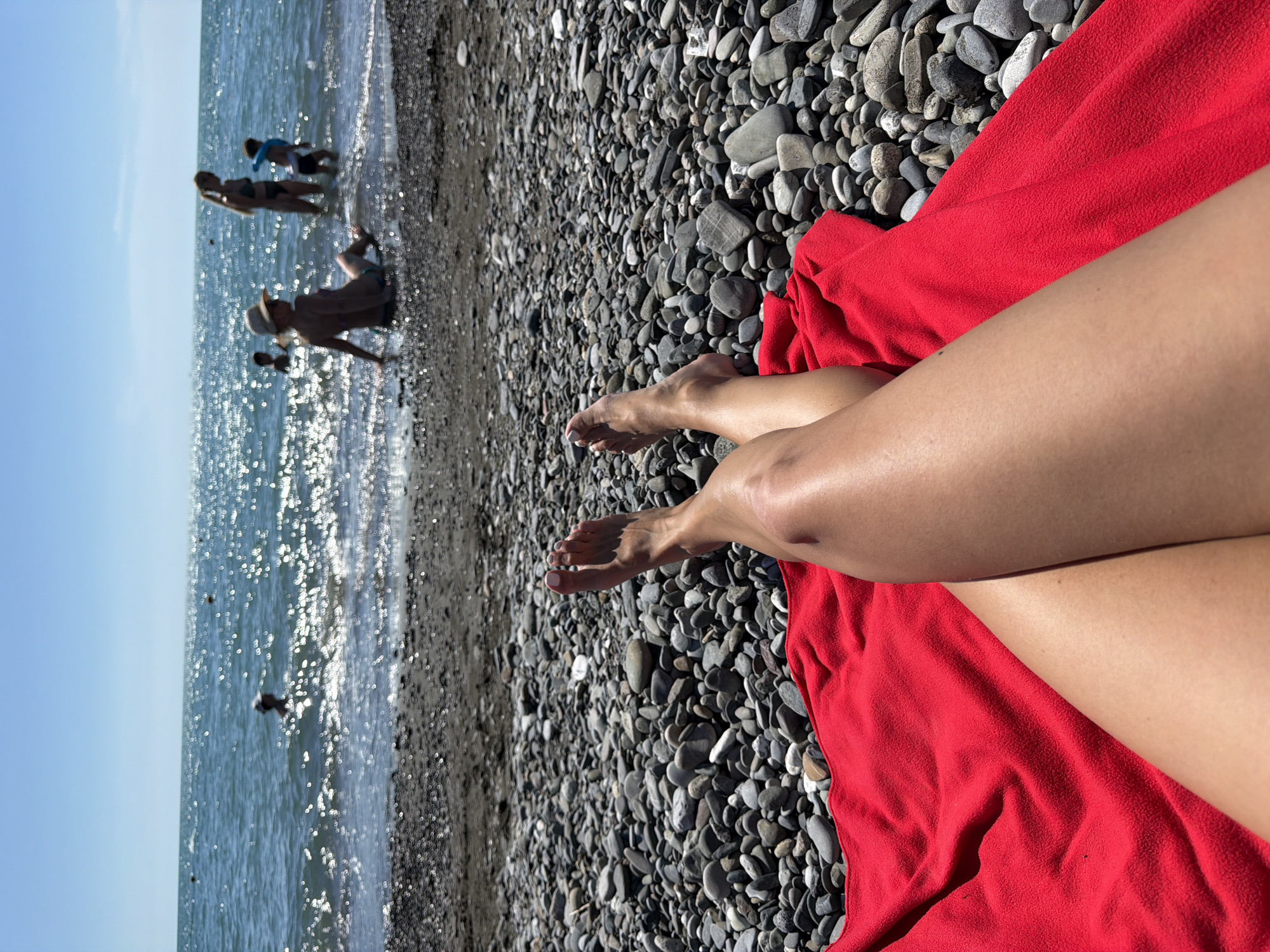 The image depicts a sunny day at the beach in Sochi, Russia. The foreground is dominated by a person's legs, which are tanned and resting on a vibrant red towel. The towel itself lies on a bed of smooth, dark grey pebbles, characteristic of many Black Sea beaches. Beyond the pebbles, the shallow, clear turquoise water shimmers intensely under the bright sun, creating a dazzling effect.
In the water, several other people are visible, appearing to be enjoying a swim or wading. One person, possibly a child, is visible further out in the water. Nearer to the shore, another person is lying down on the pebbles. The scene suggests a leisurely and relaxing day at the beach, with the sound of gentle waves and the warmth of the sun contributing to a tranquil atmosphere. The sky is a clear, bright blue, indicating fair weather and optimal conditions for beach activities. The time of day appears to be midday, given the bright sunlight and the strong reflections on the water. There is no visible text in the image.