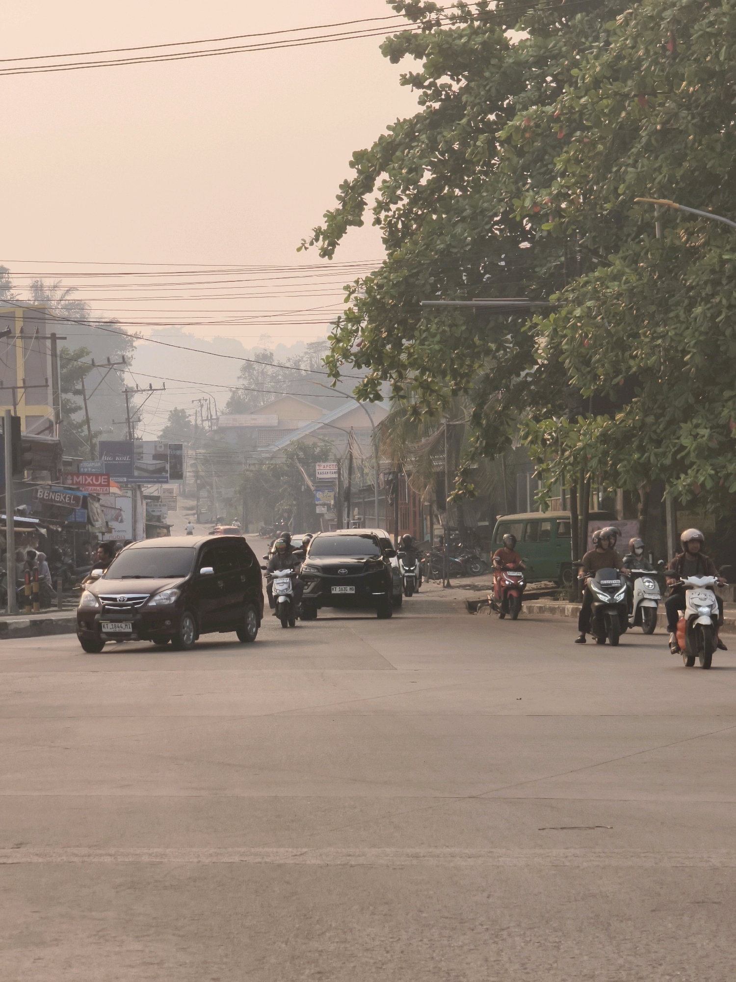 The image depicts a street scene in Samarinda, Indonesia, characterized by a slight haze in the atmosphere. In the foreground, a two-lane asphalt road is visible, with a reddish-brown car proceeding in the left lane and a black SUV following behind. Several motorcycles are also present on the road, with riders wearing helmets. To the left of the road, there are buildings with various signs, including "MIXUE" and "BENGKEL," indicating commercial establishments. Power lines are strung overhead. On the right side of the road, a large tree with green foliage dominates the scene, partially obscuring a street lamp. In the background, more buildings and trees can be seen, with hills or mountains forming the distant horizon. The overall impression is of a bustling urban environment in the early morning or late afternoon, given the soft, diffused lighting.