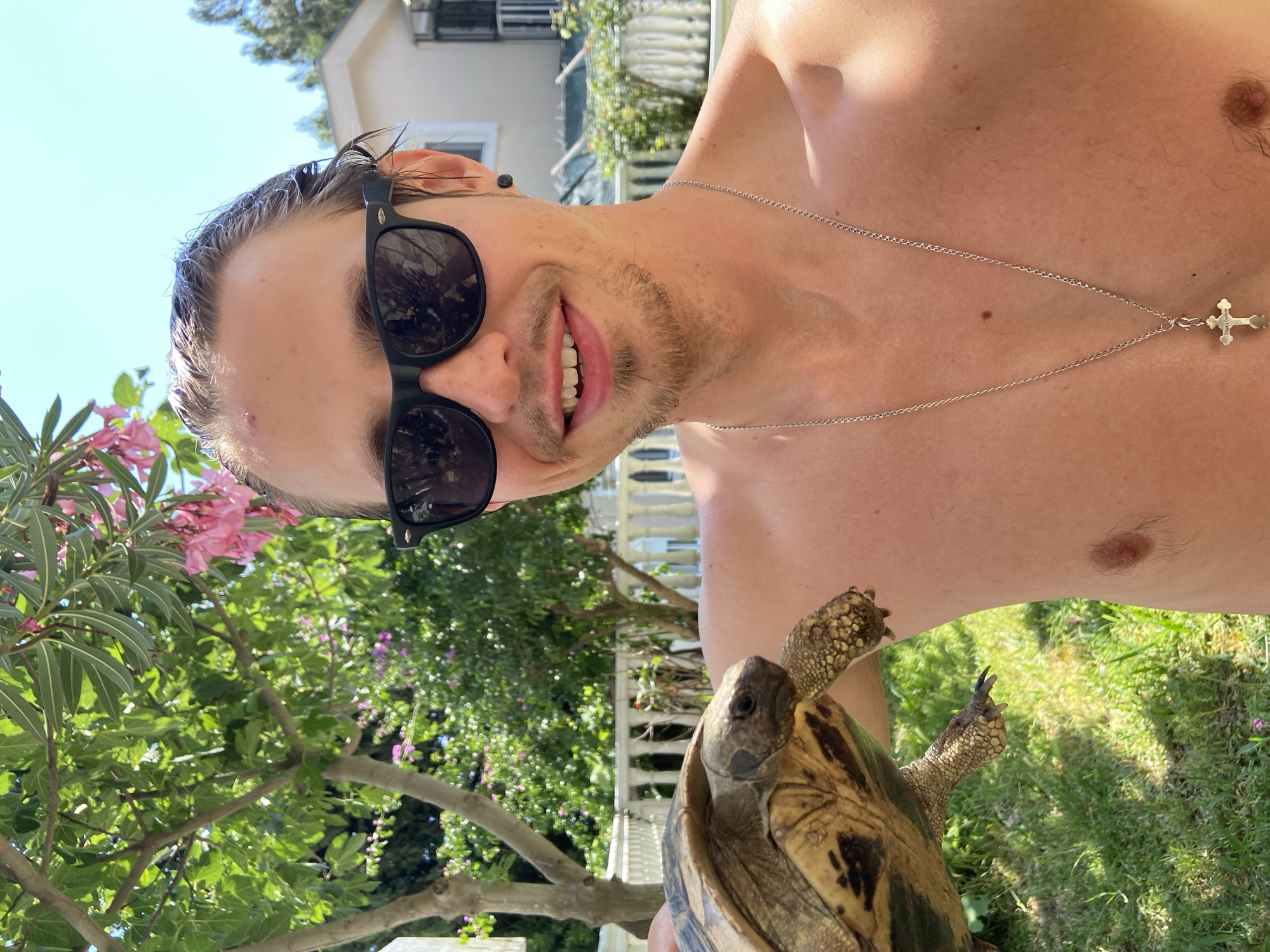 A shirtless man wearing black sunglasses and a silver cross necklace smiles at the camera while holding a tortoise. The background shows a residential area with trees, a white fence, and a building. The location appears to be in Šušanj, Montenegro, based on the distinctive architecture and vegetation.