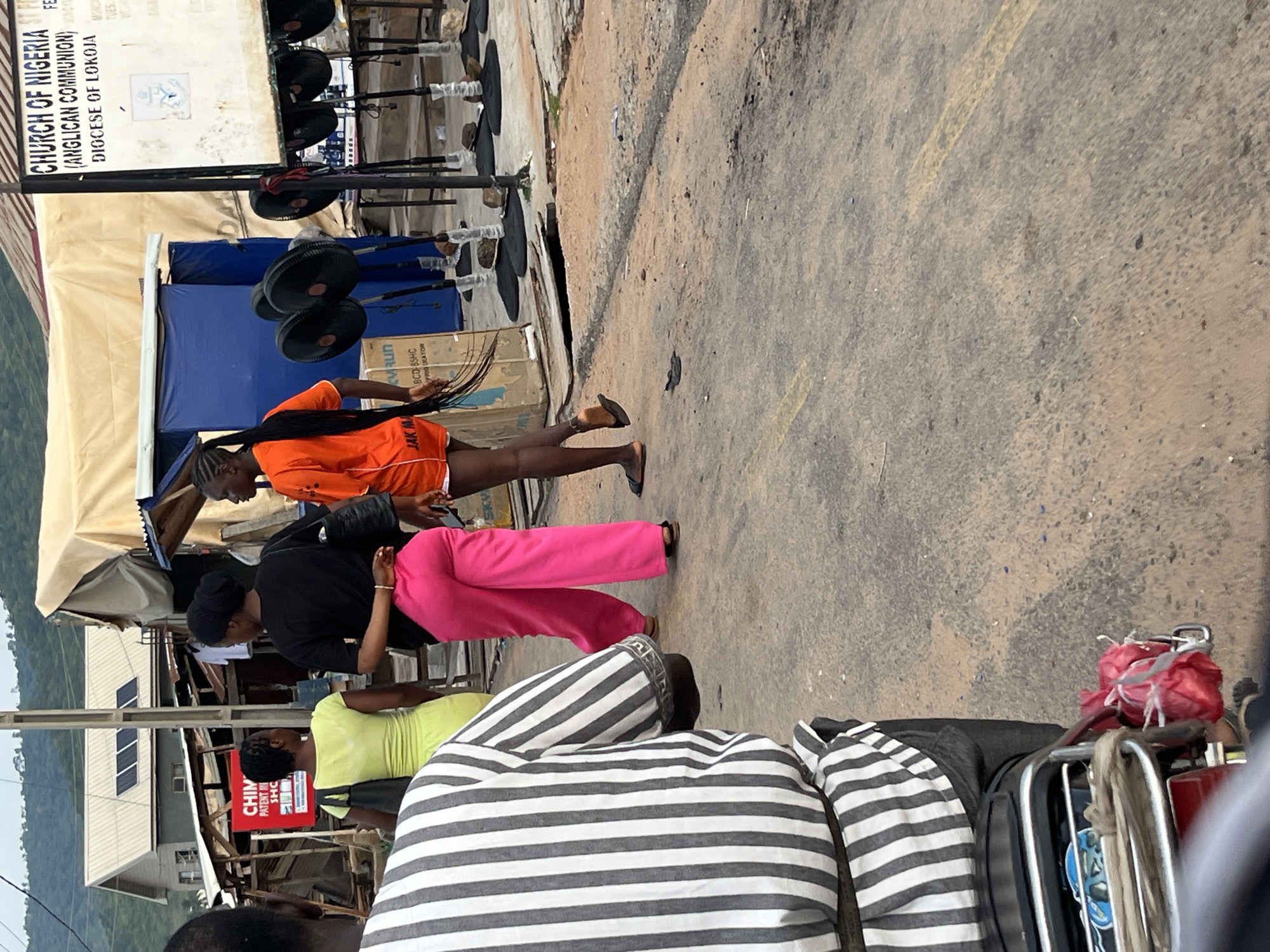 The image shows a street scene in an unknown city. In the foreground, a person wearing a black and white striped shirt is seated on a motorcycle, with the back of their head and shoulders visible. The motorcycle's rear rack has two red, bundled items tied to it. To the left of the motorcycle, a person in a lime green dress walks away from the camera, carrying a black bag. To their right, two other individuals are walking away. One is wearing a black top and bright pink pants, and the other is wearing an orange top and shorts. Behind them, a sign for the "CHURCH OF NIGERIA (ANGLICAN COMMUNION) DIOCESE OF LOKOJA" is visible. There are several fans on stands in front of a structure with a beige tarp roof. The ground is paved with asphalt and appears worn. The background shows buildings and a hillside.