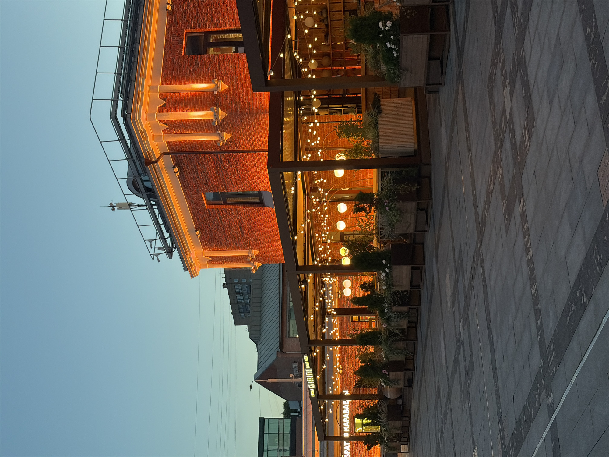 The image shows an outdoor seating area of a restaurant in Moscow, Russia, at dusk. The restaurant, likely a cafe or eatery, is housed in a red brick building with illuminated pillars and decorative lighting. String lights with round bulbs are hung above the seating area, casting a warm glow. Planters filled with greenery and flowers are arranged along the perimeter of the outdoor space. The ground is paved with large, grey square tiles, forming a patterned walkway. In the background, other buildings are visible, including a multi-story structure with windows. The sky is a clear, pale blue, typical of twilight. There are no people visible in the scene. The overall atmosphere is calm and inviting, suggesting a pleasant dining experience.