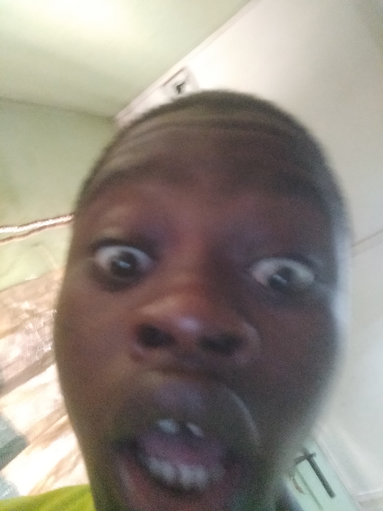 A close-up photograph shows a person's face with an exaggerated expression. The person has dark skin and appears to be making a surprised or comical face, with wide eyes and an open mouth revealing teeth. The background is indistinct, with what appear to be walls and possibly a window or curtain. There is no way to discern the location as Shangisha, Nigeria from this image.