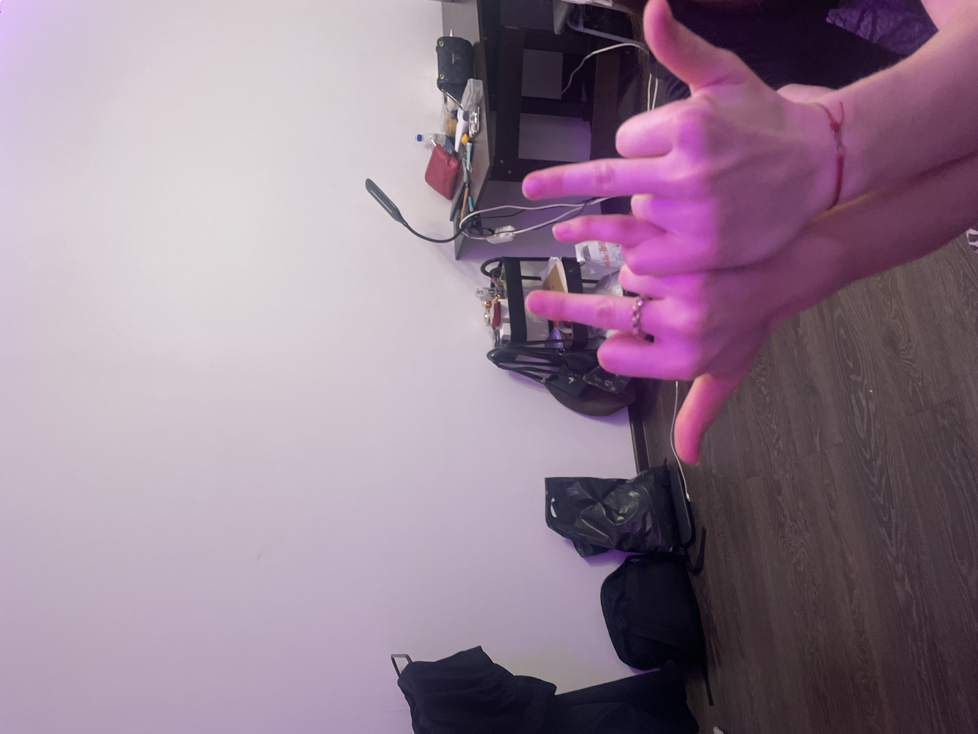 The image shows two pairs of hands, illuminated by pink lighting. One hand is in the foreground, with fingers extended in a gesture, and a ring is visible on one finger. Another hand is positioned behind it, also with fingers extended. A red bracelet is visible on one wrist. In the background, there is a wall, a desk with various items including a lamp and bags, and a floor with a wood grain pattern. The context of Sinyavino, Russia is not visually discernible from the image.