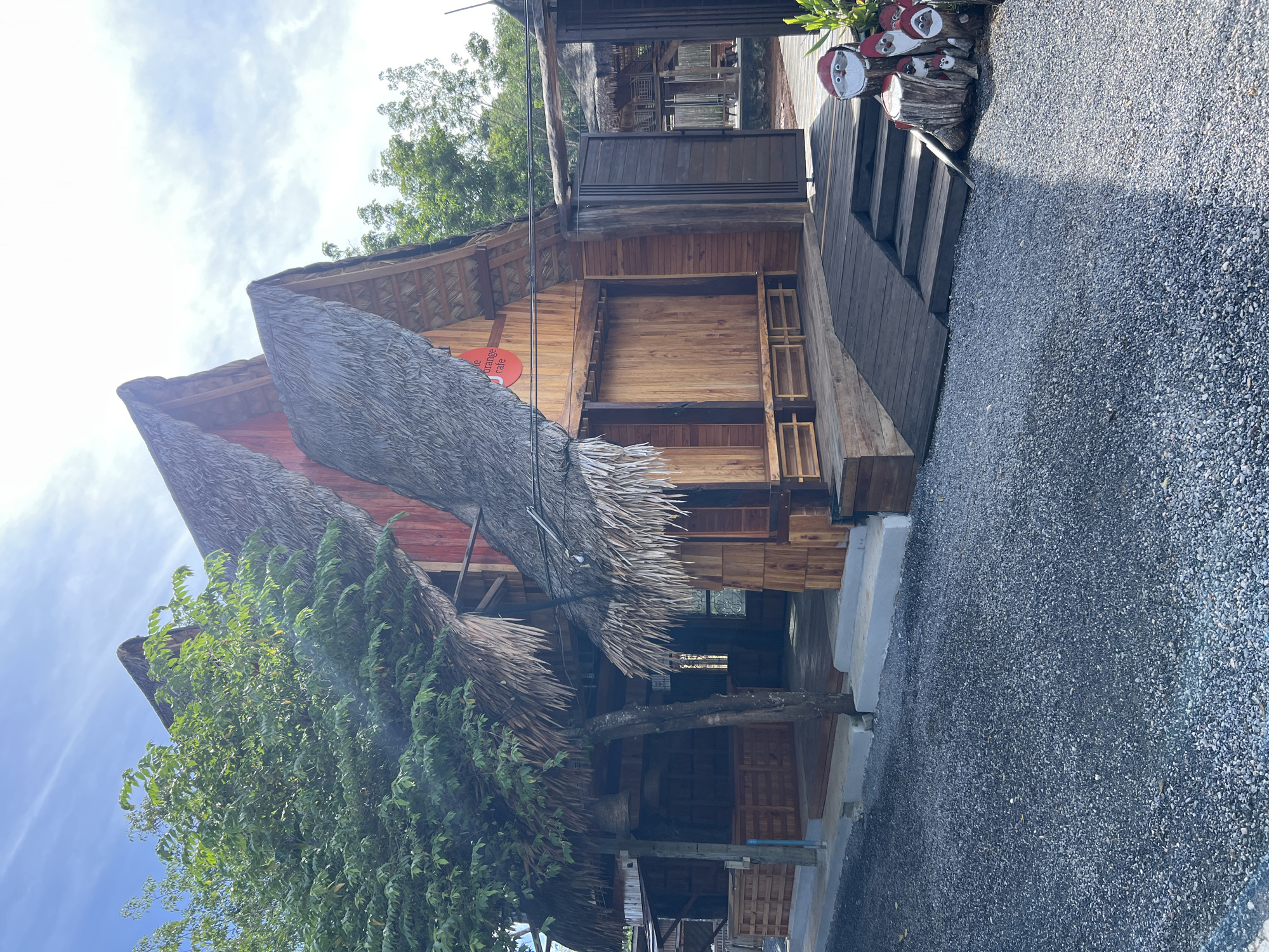 This media file depicts a rustic, open-air building in Tambon Pa Kae Bo Hin, Thailand. The structure features a traditional thatched roof made of dried palm leaves, supported by wooden beams. The walls are constructed from rough-hewn wooden planks, giving it a natural and slightly weathered appearance. In front of the building, there is a pathway covered with dark gray gravel.
To the right of the building, a wooden staircase leads up to a raised platform or deck. On this platform, there are decorative objects made from logs, painted to resemble figures, possibly gnomes or Santas, with red hats and white beards. A red circular sign with white text is visible on the side of the building.
Lush green foliage, likely trees and vines, surrounds the building, with some branches extending over and around the thatched roof. The sky above is partly cloudy, suggesting it is daytime. The scene conveys a sense of tranquility and a connection with nature.
The visible text on the red sign appears to be part of a name, possibly including the words "the orange cafe."