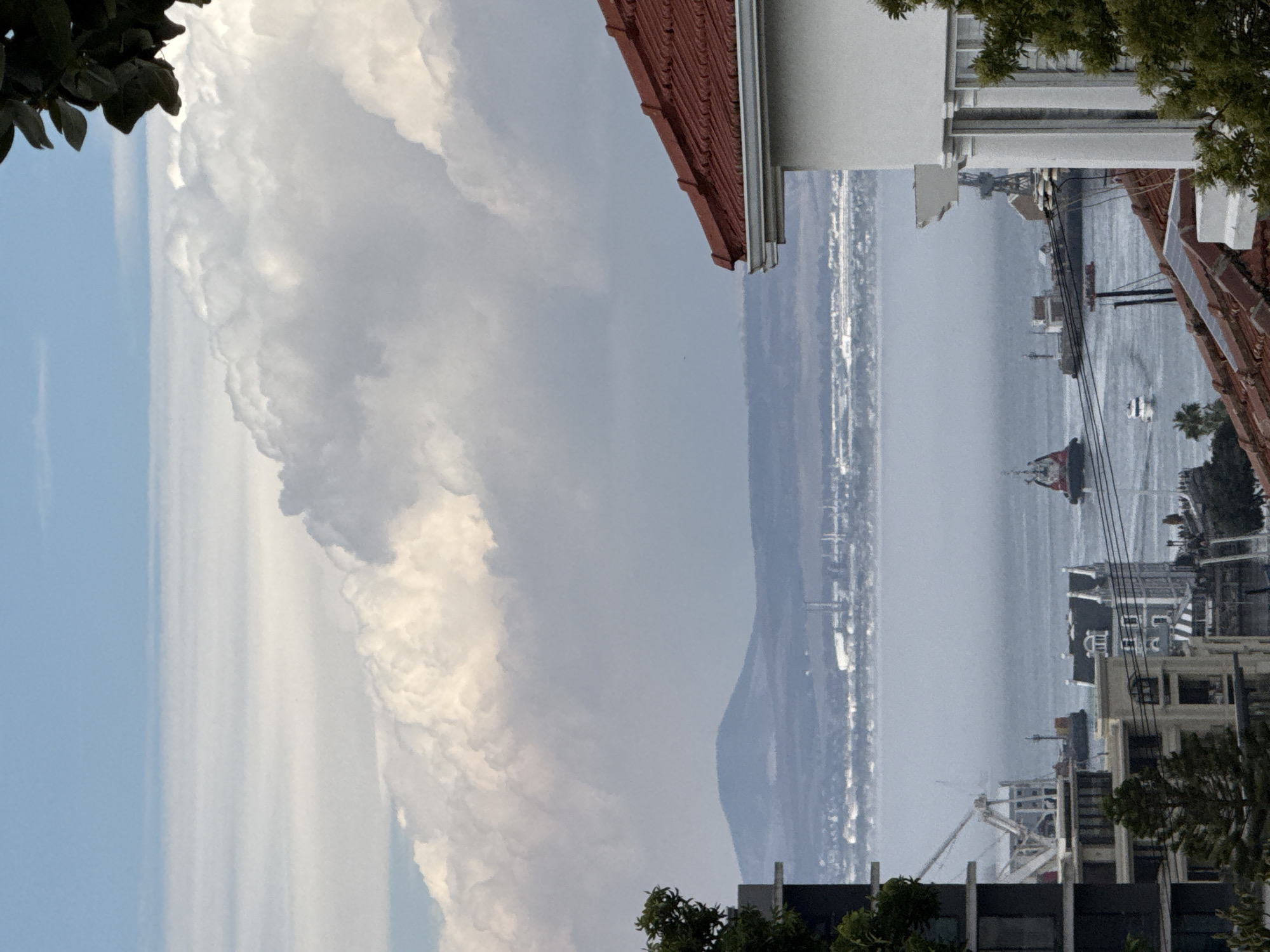 The image shows a view of Cape Town, South Africa, looking out toward the ocean. The vantage point is from a building with a red tile roof, possibly an apartment or house. The ocean is the dominant feature, appearing hazy or misty, with visible ships and boats. In the distance, there is a mountain range bordering the ocean. In the foreground, there are buildings of varying heights, typical of an urban environment. The sky is partly cloudy, with a large cumulus cloud formation. Vegetation is visible on the edges of the frame, suggesting a garden or trees in the surrounding area.