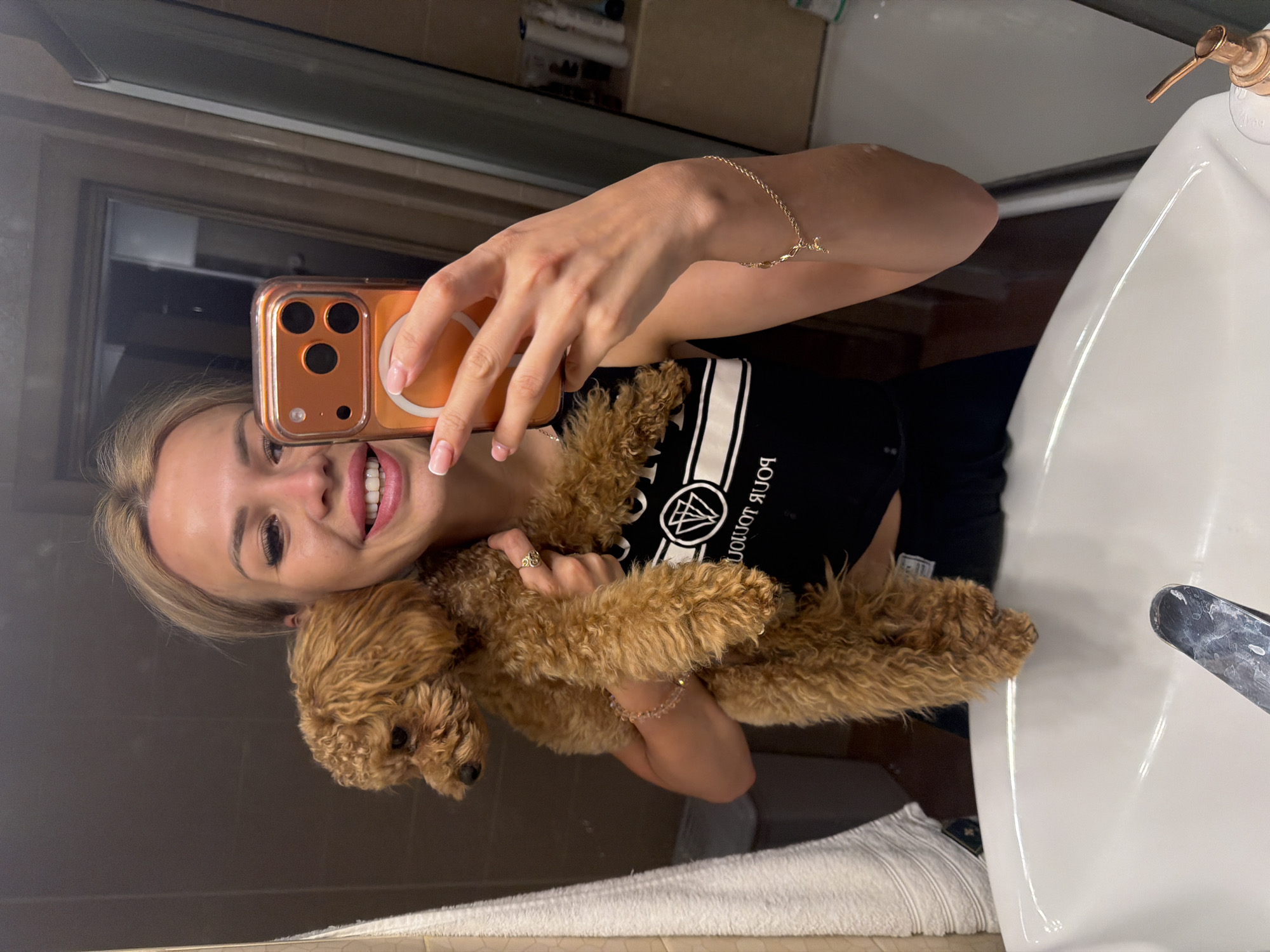 A young woman with blonde hair and bright red lipstick smiles at her phone as she takes a selfie. She is holding a small, curly brown dog in her arms. The woman is wearing a black tank top with white lettering and a delicate gold bracelet on her wrist. She also has a ring on her finger.
The setting appears to be a bathroom. A white sink and a soap dispenser are visible to the right of the woman. A mirror is behind her, reflecting the scene. A white towel is draped over the edge of the sink. The lighting suggests it could be daytime. The woman's expression is happy and playful, and the dog seems content in her arms. The image was taken in Kyiv, Ukraine. The text on the woman's shirt is partially visible and appears to be in a foreign language.