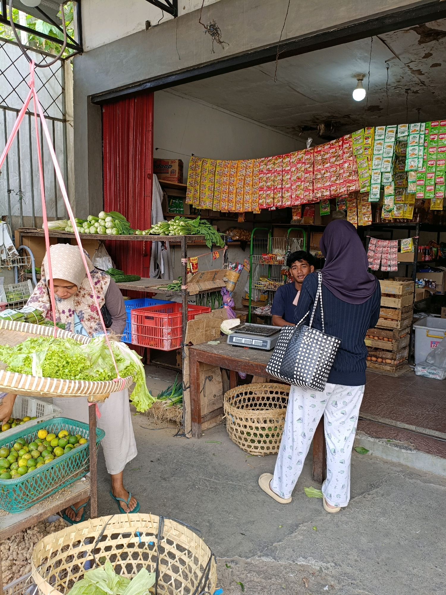 In Maguwoharjo, Indonesia, a street-side market stall displays an assortment of fresh produce and packaged goods. Two women are present, one browsing a selection of lettuce from a hanging basket, while another stands near a scale, facing away from the camera. A young man, presumably the vendor, sits behind the counter, looking towards the women. The stall is stocked with various vegetables, including limes and what appear to be ginger roots, alongside numerous brightly colored packets of packaged food products hanging from above. The scene is set against the backdrop of a modest building with a corrugated metal roof and red roll-up shutters. The ground is paved concrete.