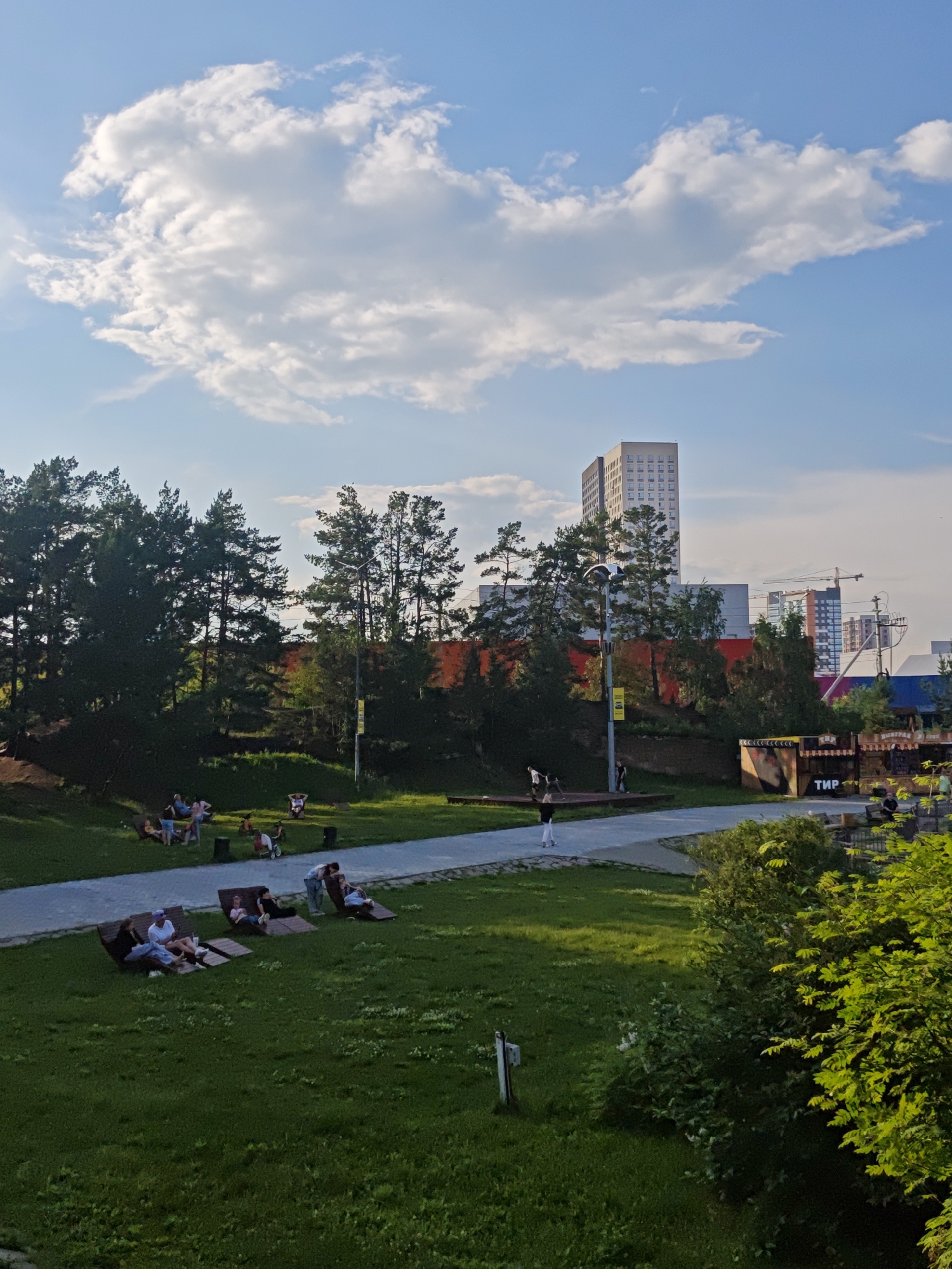 The image captures a sunlit park scene in Yekaterinburg, Russia, possibly during the late afternoon or early evening, given the warm lighting and long shadows. The sky is a vibrant blue, dotted with fluffy white clouds, suggesting fair weather.
In the foreground, a lush green lawn is populated by several groups of people relaxing on lounge chairs and blankets. One group of three individuals sits on lounge chairs, engaged in conversation. Another individual reclines on a lounge chair, looking away from the camera. Further back, a group of four people is gathered near some trees, and another individual is seated on a bench.
A paved pathway curves through the park, separating the grassy areas. Along this pathway, there are scattered trees providing shade. In the middle ground, a collection of food stalls or small shops with colorful facades are visible. The word "ТИР" (which translates to "shooting range" in Russian) is prominently displayed on one of these structures.
The background features a dense line of dark green trees, primarily pine trees, which create a natural backdrop. Beyond the trees, modern high-rise buildings, including a tall, light-colored skyscraper with numerous windows and cranes indicating ongoing construction, dominate the urban skyline. The scene conveys a sense of leisure and community gathering in an urban park setting.