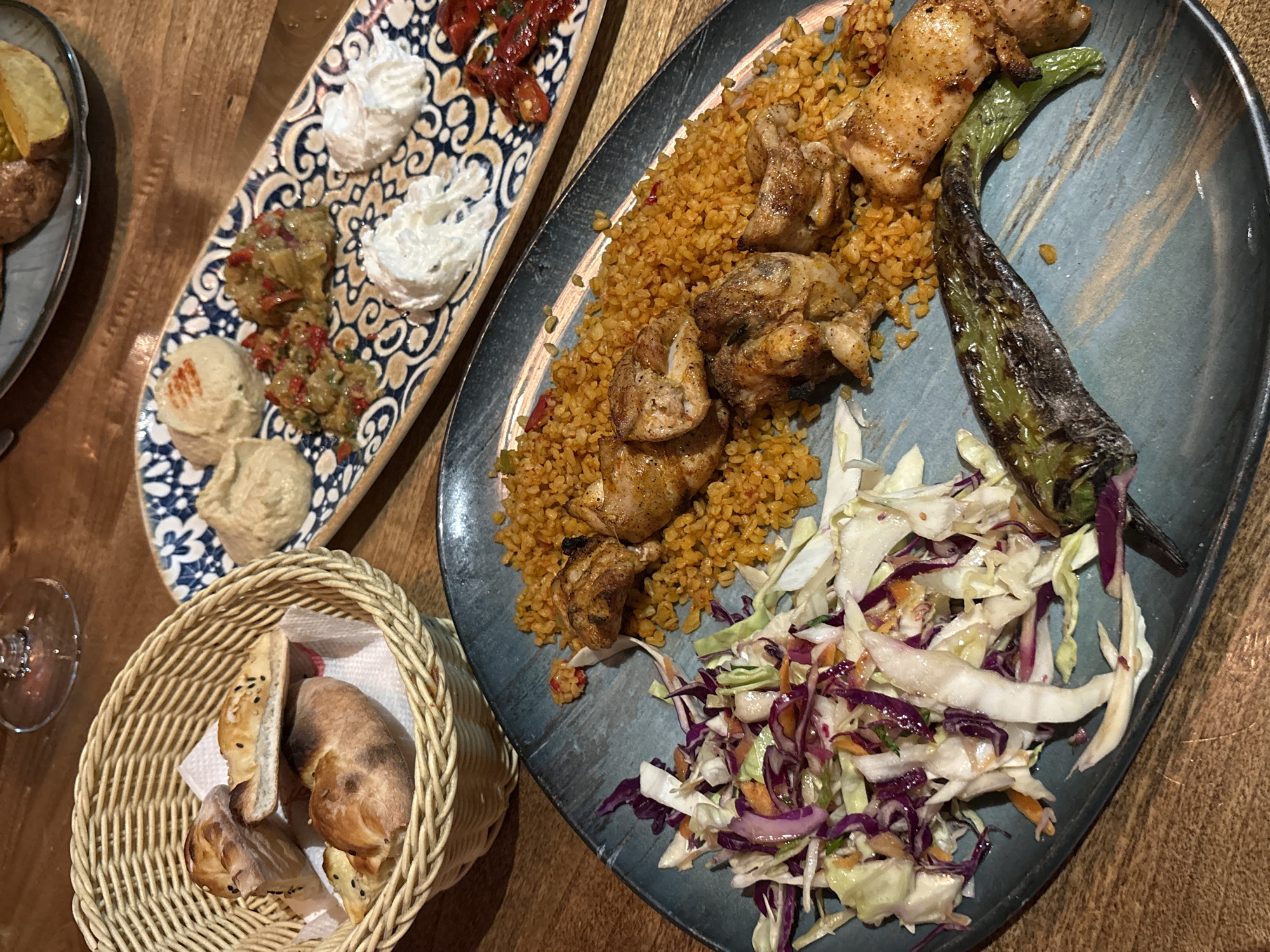 The image shows a table setting with two plates of food and a basket of bread. The plate on the right contains grilled chicken pieces arranged over a bed of bulgur with a grilled pepper and a small salad of shredded cabbage and carrots on the side. The plate on the left has several dollops of different dips: what looks like hummus, ajvar, and labneh, plus a couple of grilled tomatoes. A basket with flatbread is in the lower left corner of the image. A small glass is partially visible in the lower left, near the bread. The table is made of wood. The scene is likely a restaurant. Given the style of the food, it could possibly be located in Plovdiv, Bulgaria. No people are visible.