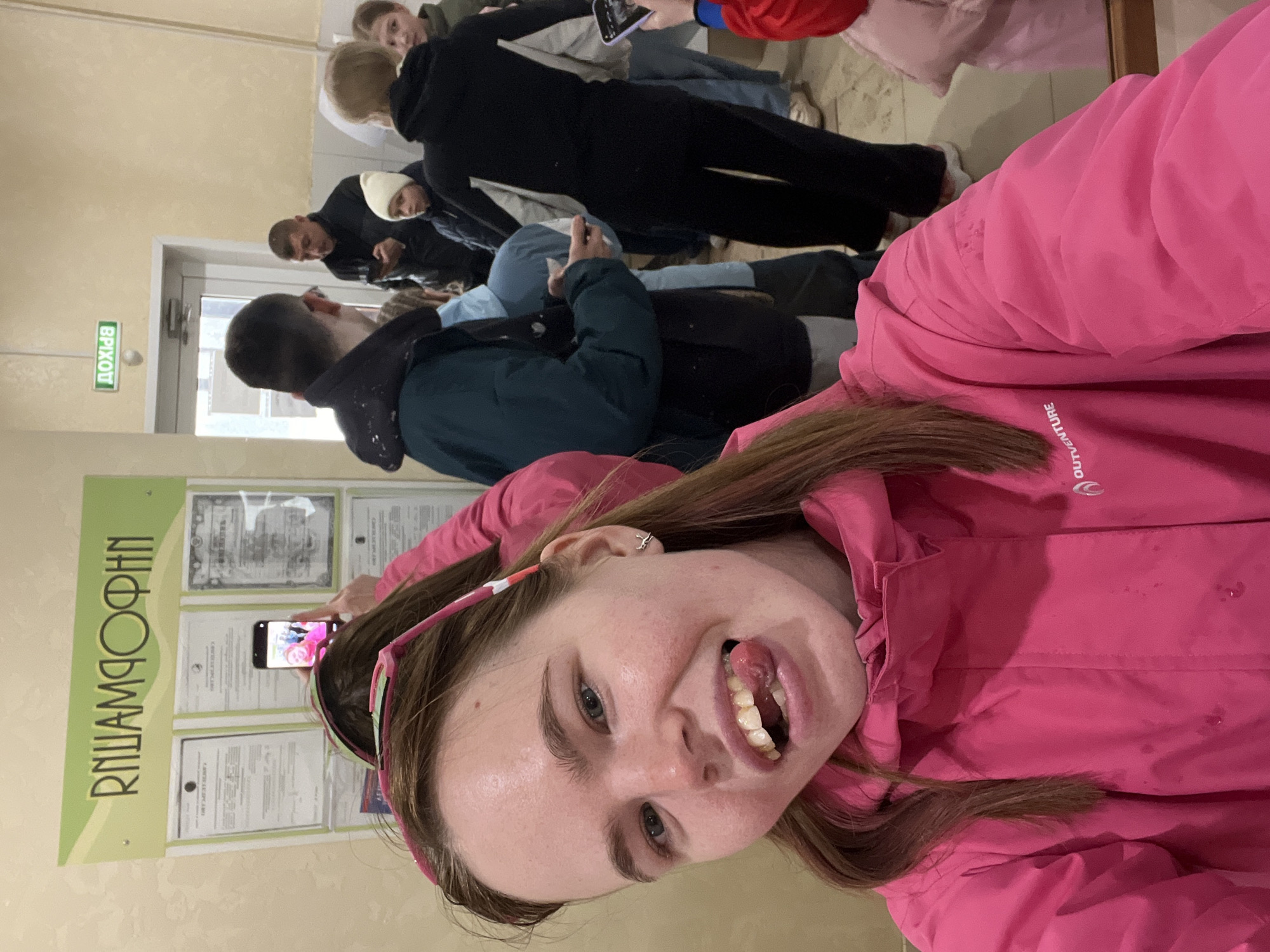 A woman in a pink jacket takes a selfie, sticking her tongue out. In the background are several people standing and sitting in what appears to be an interior space. One person is holding a phone, possibly taking a picture of a sign on the wall. The sign has Russian text and the word "ОФИН". A door with the Russian word "ВЫХОД" (Exit) is also visible. The location is likely a public building in the city of Gory, Russia.