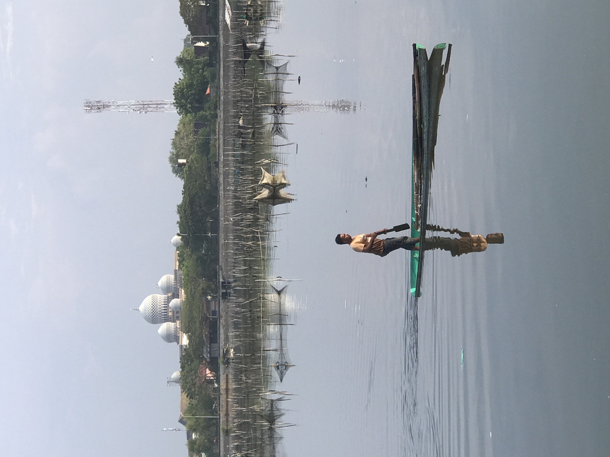 The image captures a waterfront scene in Lhokseumawe, Indonesia. In the background, there is a building with a series of white domes. To the left of this, a tall, metal lattice tower is visible. The shoreline is lined with trees and various structures, including what appear to be fishing traps or weirs extending into the water. In the foreground, a man stands on a makeshift raft or small boat in the water, using a paddle. The water is mostly calm, reflecting the sky and the structures on the shore.