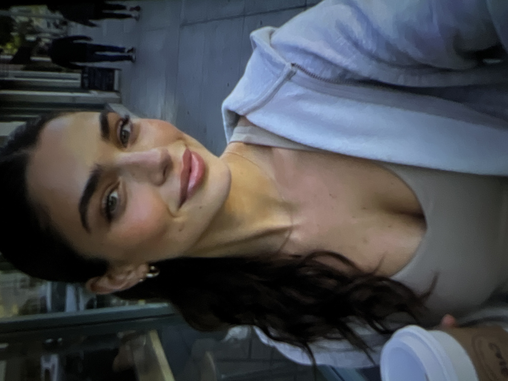 A young woman with dark hair pulled back in a ponytail and wearing a light-colored hooded jacket and a tank top smiles as she holds a coffee cup in her left hand. Her right hand is not visible. She is standing on a sidewalk next to a street with other pedestrians visible in the background. The environment appears to be an urban setting with buildings lining the street. There is no visual information within the image to suggest the location is Pyatigorsk, Russia.