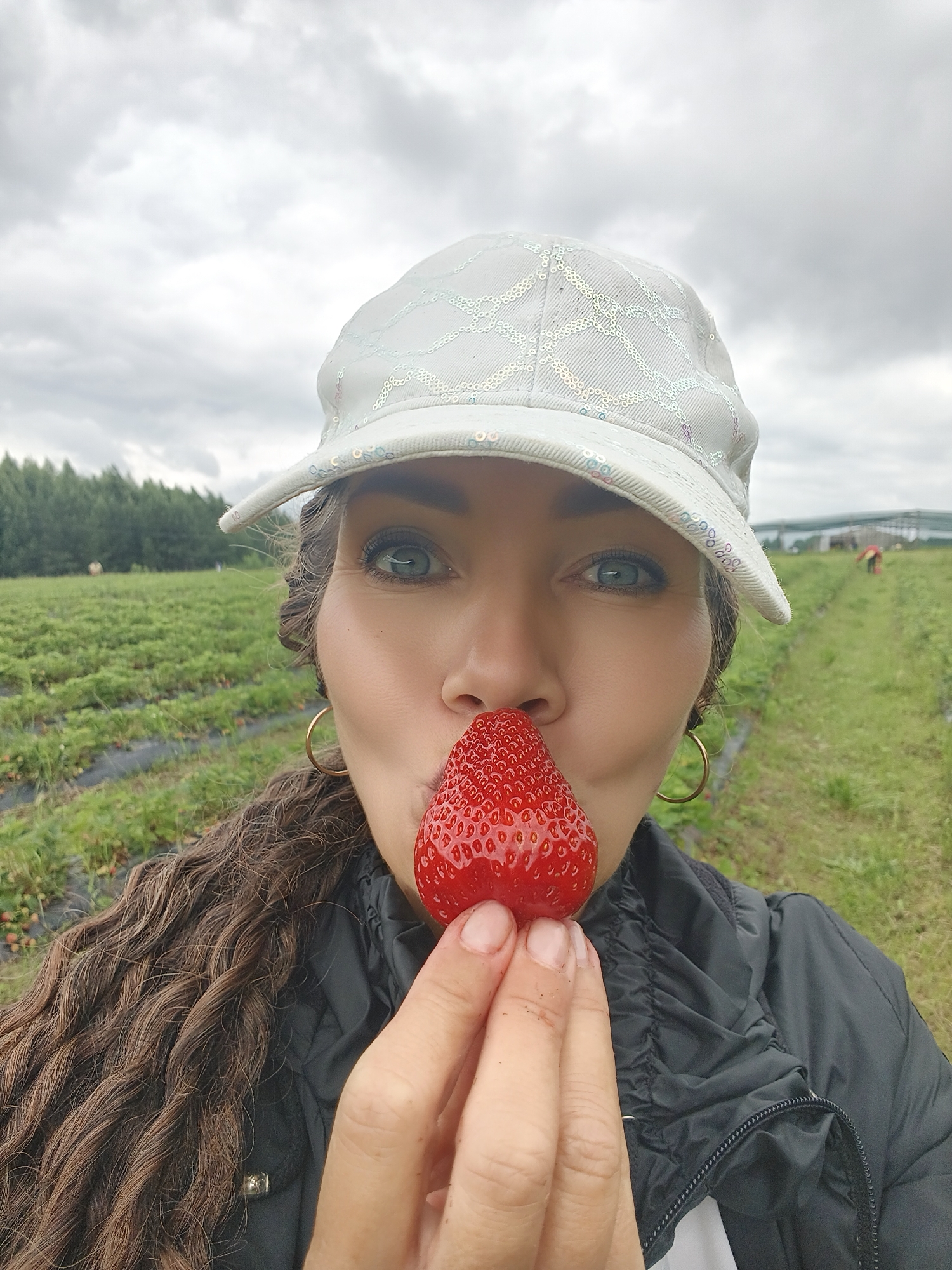 A woman in a white sequined baseball cap and black jacket holds a strawberry up to her face, puckering her lips. She has long, wavy brown hair and is wearing large gold hoop earrings. The background is a strawberry field with rows of plants under a cloudy sky. Two people are visible in the distance, appearing to be picking strawberries.