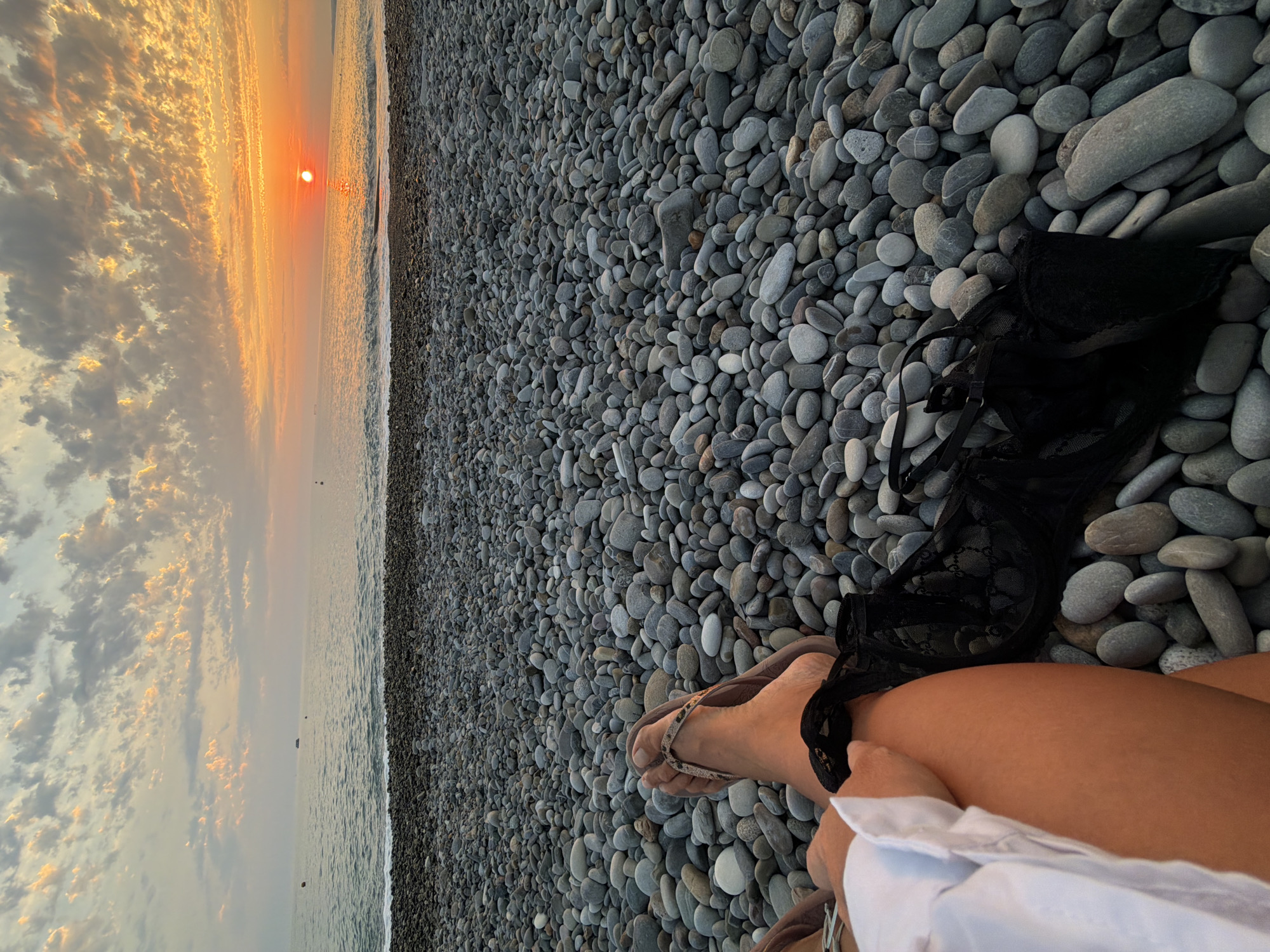 The image depicts a beach in Shepsi, Russia, at sunset. The foreground is dominated by a dense scattering of grey and beige pebbles. A person's legs and feet, wearing sandals, are visible on the left side of the frame. A black lace bra lies on the pebbles next to their legs. In the midground, the sea gently laps at the pebble shore. The background features a wide expanse of the sea under a sky filled with dramatic clouds illuminated by the setting sun. The sun is a bright orange orb low on the horizon, casting a warm glow over the water and the sky.