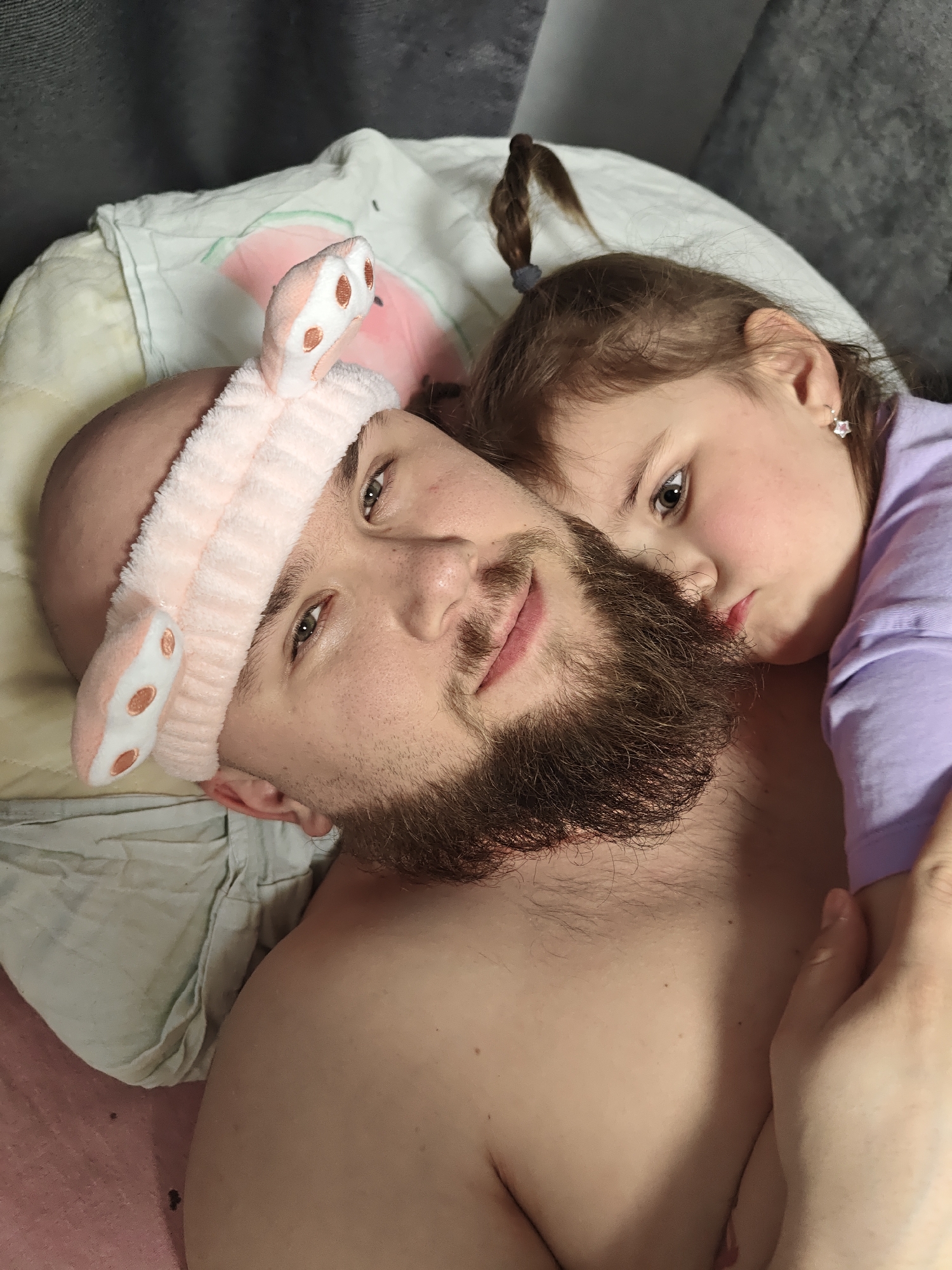 This close-up portrait, taken in Chaikovsky, Russia, captures an intimate moment between a man and a young girl lying down together.
The man, with a bald head and a prominent dark brown beard, is looking directly at the camera with a gentle expression and green/hazel eyes. He is wearing a whimsical light pink fuzzy headband with white and brown spotted bunny ears, positioned on his head. His upper torso is bare, revealing skin without a shirt.
Nestled close to him, resting her head on his shoulder or chest, is a young girl. She has light brown hair, with a small braid secured by a dark elastic visible near the top of her head. Her visible eye (appearing dark or brown) is also directed generally towards the camera, though her gaze is softer and less direct than the man's. She wears a light purple long-sleeved shirt, and a small, delicate star-shaped earring is visible in her right ear. A hand, likely hers, is partially visible resting on the man's chest.
They appear to be on a bed or couch, with a white pillow featuring a faint pink design behind the man's head. A textured grey fabric, possibly upholstery or a headboard, is in the background. Another light-colored, possibly green, fabric is visible crumpled near the man's left shoulder. The overall mood is affectionate, peaceful, and relaxed. There is no visible text in the image.