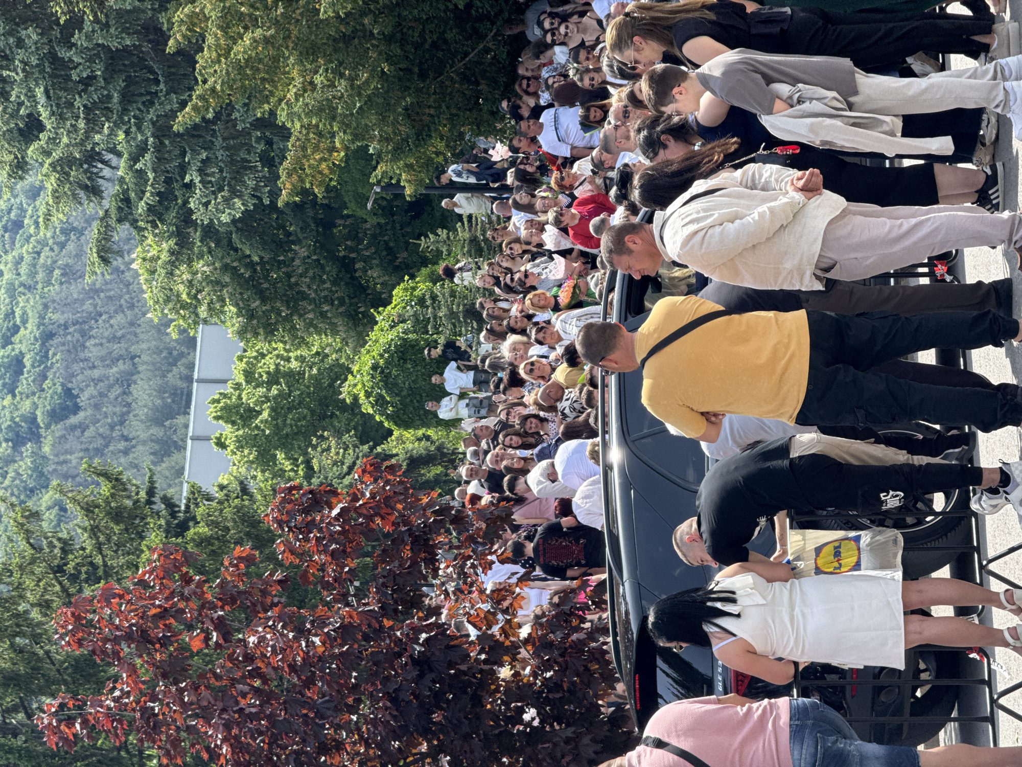 A crowd of people has gathered in Shumen, Bulgaria, possibly for a public event or demonstration, as can be discerned from the density of the gathering. The crowd extends into the distance, filling the available space.
Some individuals at the front of the crowd are standing near a black vehicle and a metal barrier. The people visible at the front seem focused on something beyond the immediate foreground, as their gazes are directed towards the center of the gathering.
The environment consists of urban landscaping with trees and vegetation surrounding the gathering. One tree has distinct red leaves, contrasting with the green foliage of the other trees, providing a visual separation within the landscape. The day appears sunny and bright, casting shadows on the ground and the crowd.