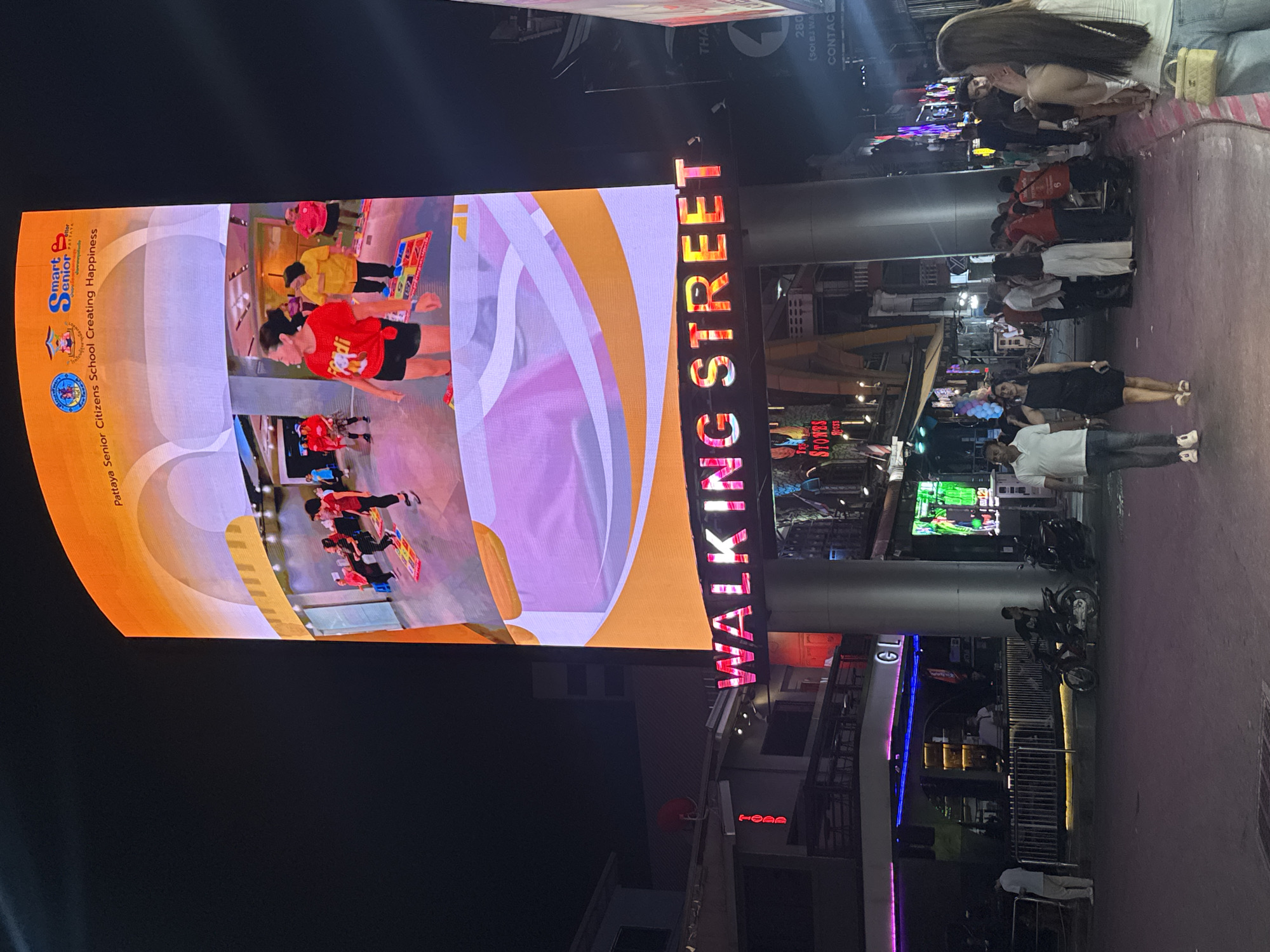 The nighttime photograph captures a vibrant street scene in Pattaya City, Thailand, dominated by a large vertical LED screen and a prominent "WALKING STREET" sign. The LED screen, positioned vertically on the left side of the image, displays an indoor activity involving several people, likely senior citizens, participating in a game or exercise on colorful mats. Text on the screen reads "Pattaya Senior Citizens School Creating Happiness" and includes a "Smart Senior" logo, suggesting an educational or recreational program. One woman on the screen wears a red shirt with "Fendi" visible.
To the right of the screen, the illuminated red letters clearly spell out "WALKING STREET". Below this sign and extending into the background, the street is bustling with numerous people walking and standing, indicative of a lively evening atmosphere. Some individuals are seen near parked or moving motorbikes. Buildings line the street, featuring various lights and signs, including a partially visible sign for "STONES HOUSE". Other visible text fragments include "TODD" and "280 (SOLBJ WA CONTAC". The overall scene depicts a busy urban environment at night, typical of a well-known entertainment district, with a focus on both local community activities displayed on the screen and the general public going about their evening on the street.
