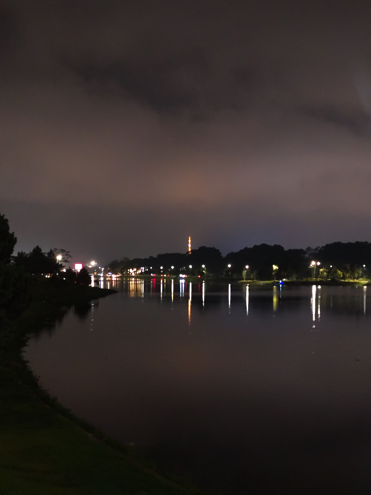 The image depicts a nighttime scene at Xuan Huong Lake in Da Lat, Vietnam. The lake surface reflects the lights from surrounding street lamps and buildings. In the background, a tall structure, likely a radio tower or communications tower, is illuminated. The sky is overcast, with clouds obscuring the upper portion of the image. Along the lake's edge, there is vegetation, including trees and grass. There are no people visible in the image. The scene is illuminated by artificial lights, creating reflections on the water.