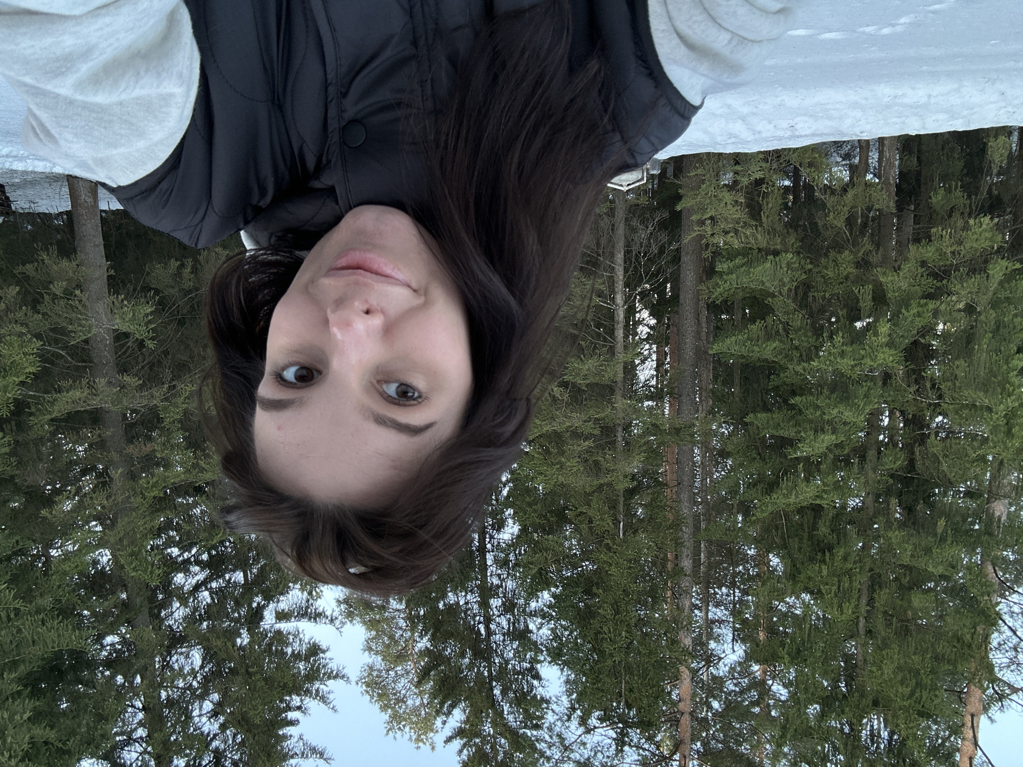 An outdoor selfie captured in Прикамский, Russia, depicts an adult female positioned against a snowy forest backdrop. The subject, with long dark brown hair and brown eyes, is looking directly at the camera with a slight smile. She is wearing a black quilted puffer vest over a light-colored long-sleeved garment, likely white or light grey. The foreground consists of an even layer of white snow. Behind the subject, a dense array of tall evergreen trees with dark green needles and visible brown trunks rises towards a clear, light blue sky. Natural daylight illuminates the scene.