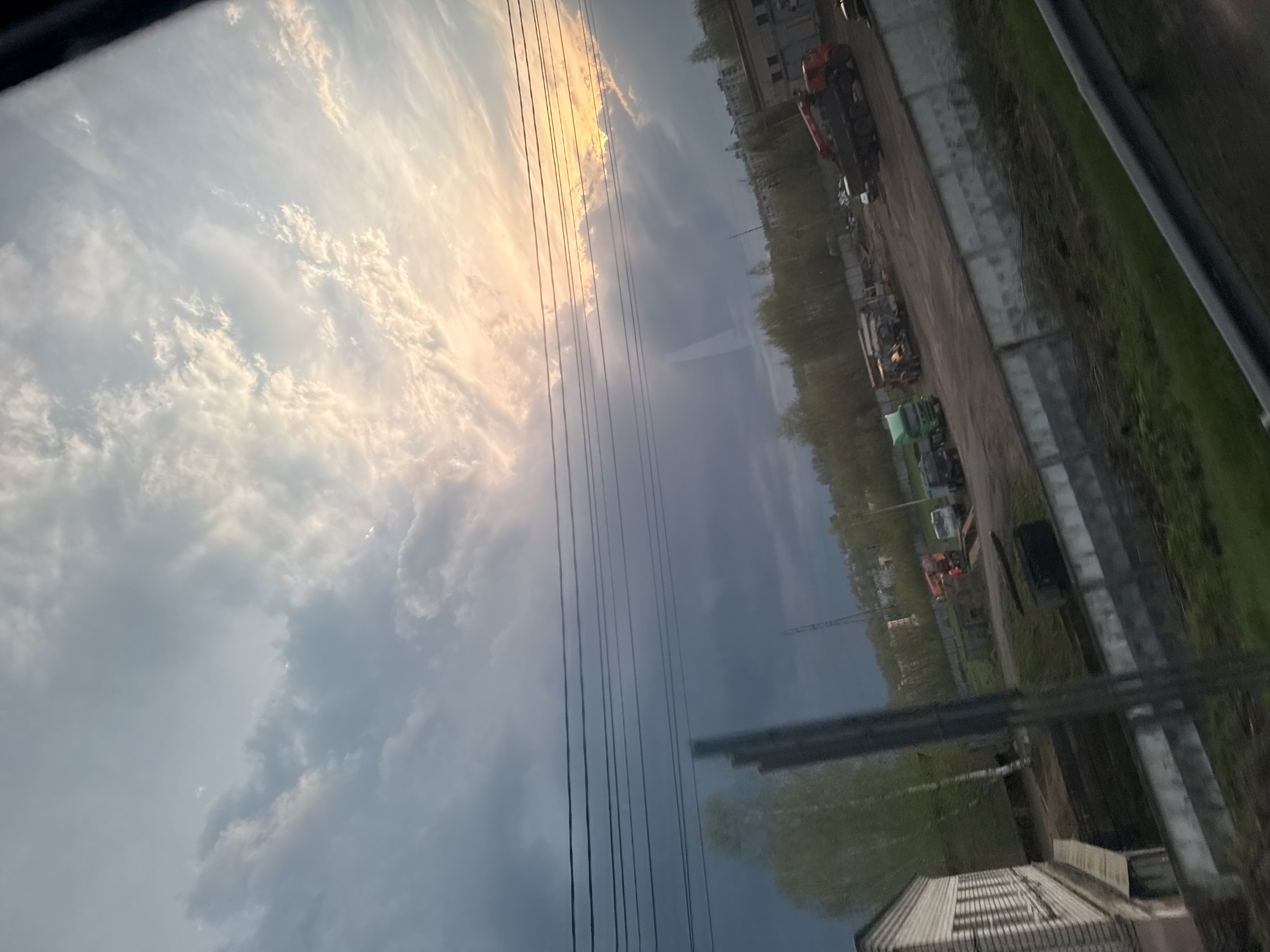The image shows a view from an elevated position in Yakhroma, Russia. The sky is cloudy with a mix of dark and light patches, suggesting a transitioning weather pattern. There are power lines visible in the foreground, obscuring parts of the scene.
Below, an industrial or commercial area is visible. Several vehicles, including what appears to be a truck and construction equipment, are parked. Buildings line the edge of the developed area. The image is somewhat blurry, with the scene captured at an angle.