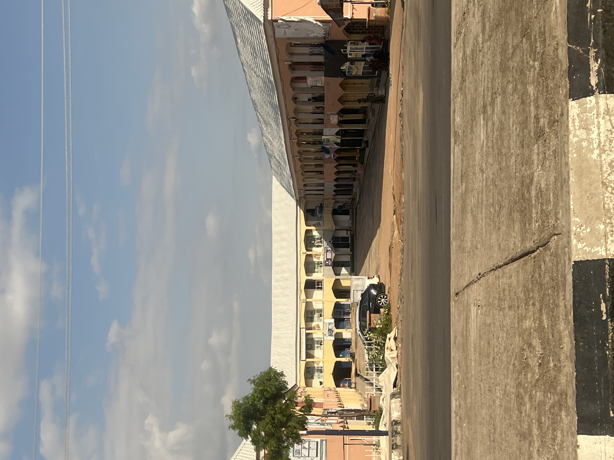 This media file depicts an everyday street scene in Howayi, Nigeria, viewed from a slightly elevated vantage point. On the left side of the image, a series of multi-story buildings line a road. The prominent building in the center features numerous units with arched balconies or open corridors, painted in warm tones of peach, pink, and yellow. Many of these balconies have clothes hanging to dry, indicating residential use. The building is topped with a light-colored corrugated metal roof.
Parallel to these buildings runs a dirt or unpaved road. A dark-colored car, possibly an SUV, is parked on the roadside, partially hidden by green shrubs and plants. A large, weathered concrete wall or barrier dominates the right foreground, displaying a distinct crack and textured surface, along with some black and white striped markings on its side.
The sky above is bright and clear blue, dotted with scattered white cumulus clouds. Several thin power lines stretch across the upper left quadrant of the sky. A large, leafy green tree stands on the left side of the road, closer to the viewer.
The scene suggests a sunny day, likely mid-morning or afternoon, given the bright illumination. No specific activity or event beyond daily life is explicitly taking place. The overall impression is a calm, ordinary street in an urban or semi-urban setting. No discernible text is visible in the image.
