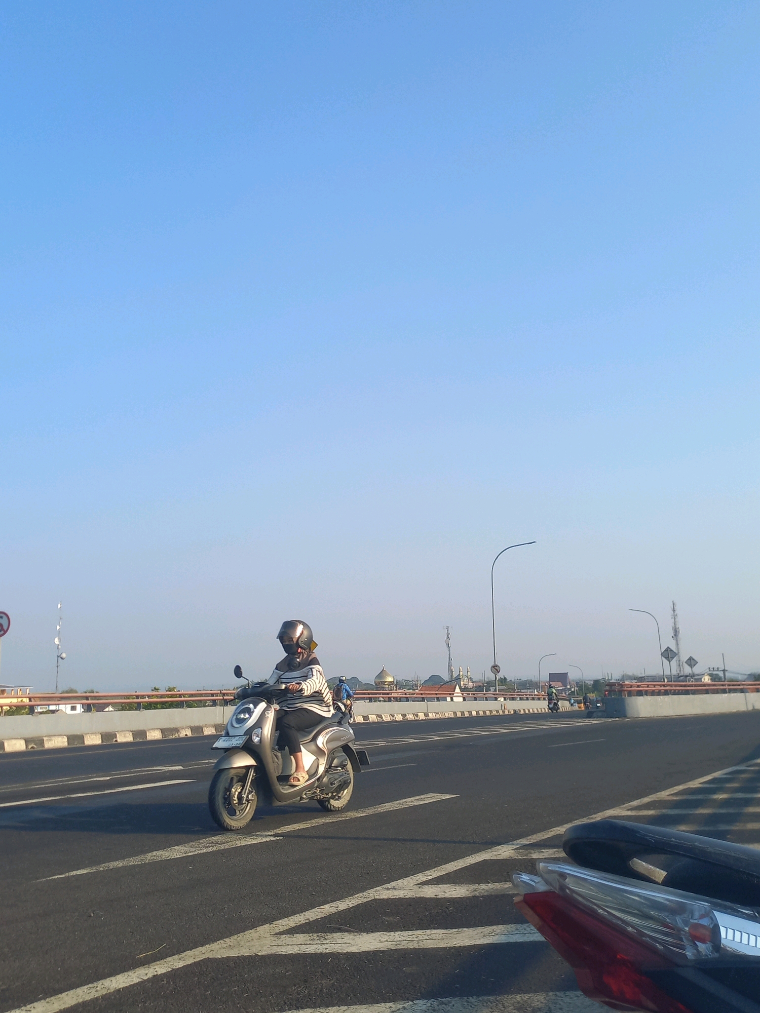 A person wearing a helmet and a striped shirt is riding a silver scooter on a road in Ploso, Indonesia. The sky is clear and blue. In the background, there are streetlights, buildings, and other vehicles on the road. The road has white markings. Part of another scooter is visible in the foreground.
