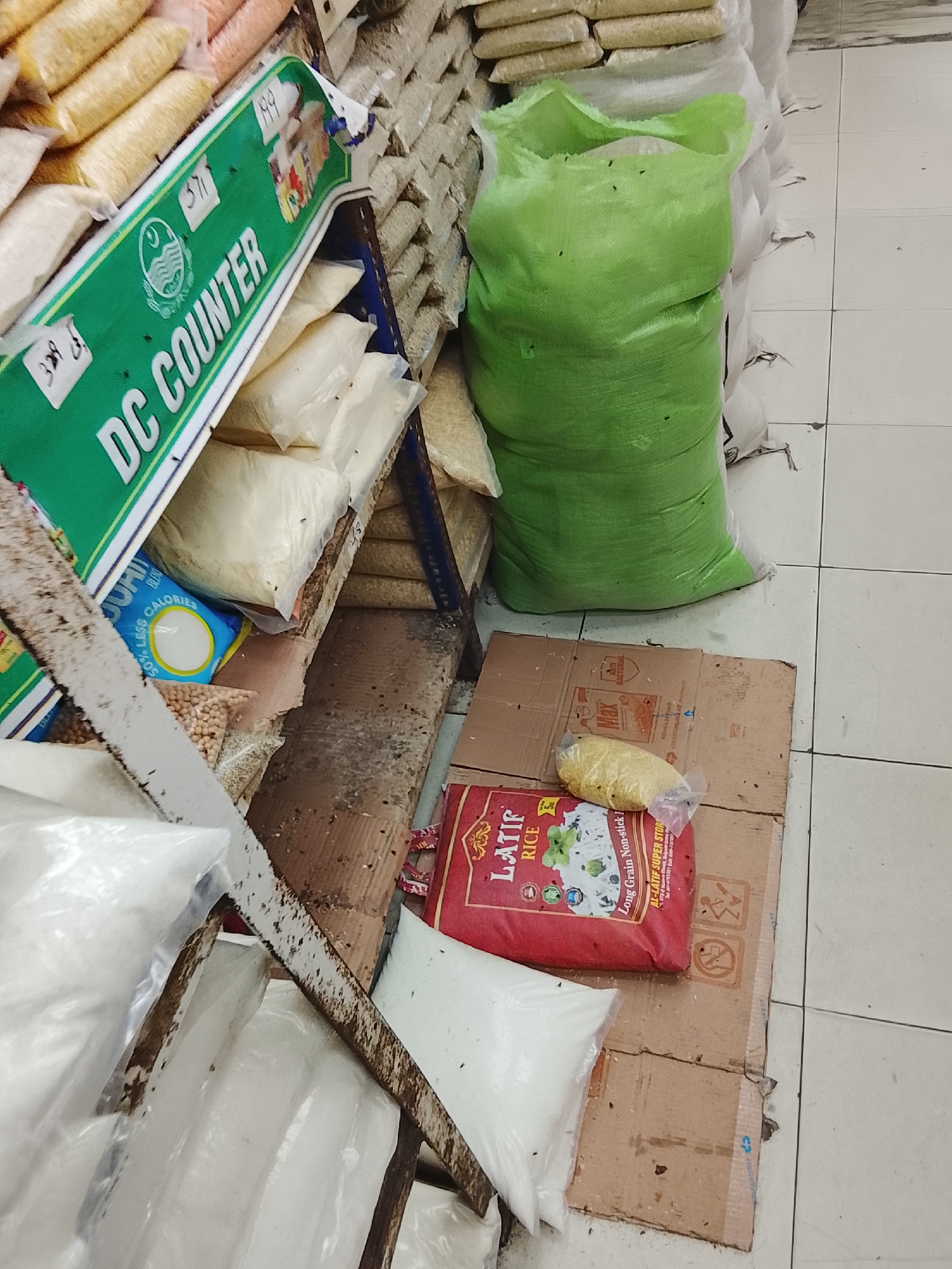 The image shows a scene within a store, possibly in the Gulgasht Colony, Pakistan, judging by the text on the Latif Rice packaging. Shelves are stocked with bags of various food items, including beans and lentils. A green bag sits on the floor next to boxes of Latif Rice and other prepackaged items. The shelves and cardboard boxes on the floor appear worn and show signs of age. The tile floor is visible, and the arrangement suggests a retail or storage area. The “DC COUNTER” sign indicates a point of sale or a dedicated section within the store. The presence of flies on several surfaces suggests sanitation may be an issue.