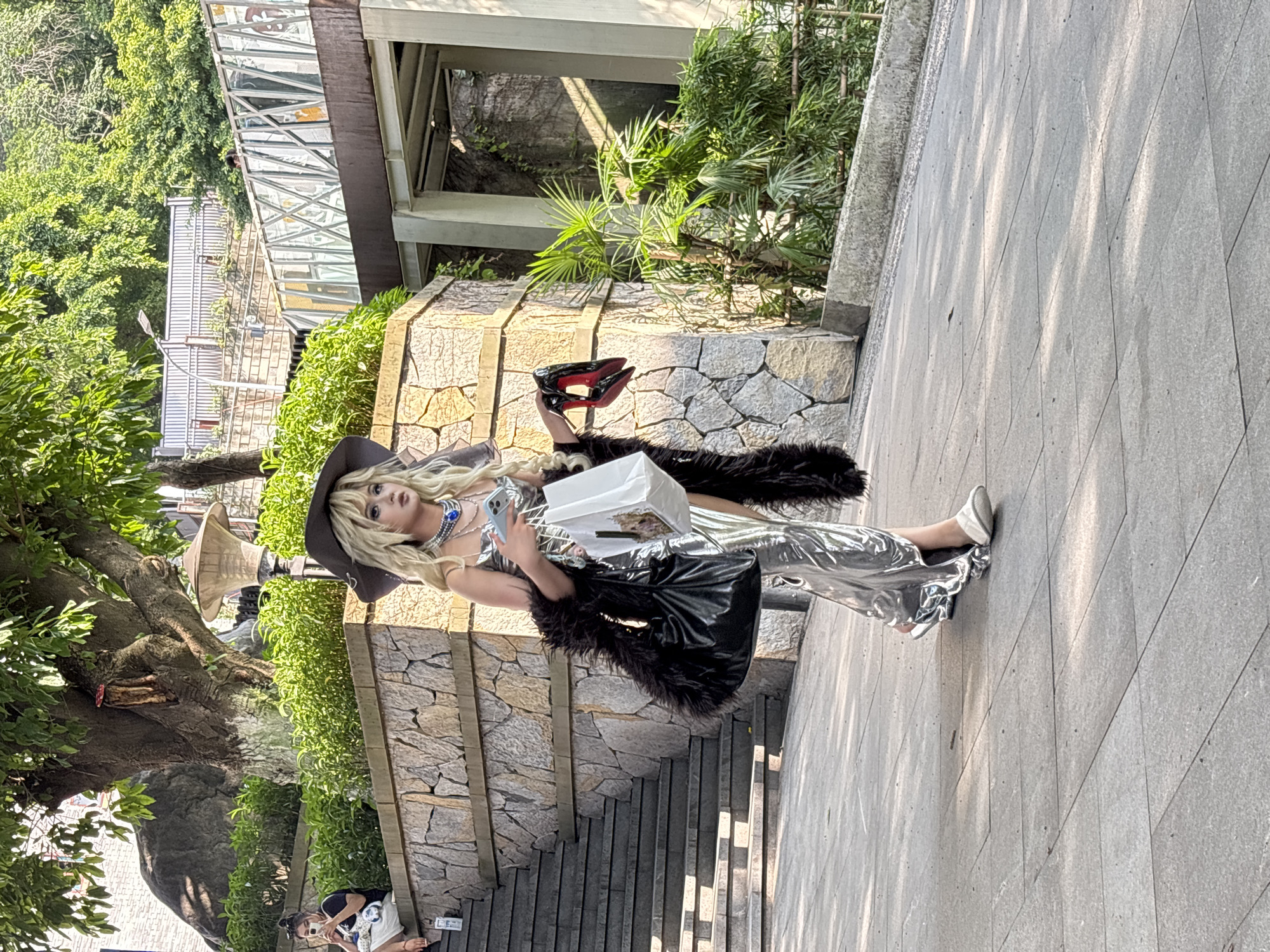A person is walking on a paved pathway in Yuzhong District, China, likely in an outdoor public space with tiered stone landscaping and lush green foliage. The individual is wearing a shimmering silver dress, a wide-brimmed hat, and a dark, fluffy wrap or scarf. They are carrying a black purse and a white shopping bag, and in their right hand, they are holding a pair of red high-heeled shoes. To the left of the main subject, a set of stone stairs is visible, with another person sitting on them and holding a mobile phone. In the background, there are trees, a stone wall, and architectural elements suggesting a multi-level structure. The lighting indicates it is daytime, with shadows cast on the pathway.