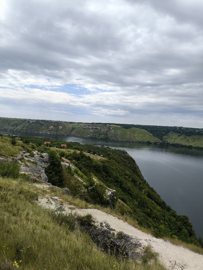 The image displays an elevated, panoramic view of a wide river valley in Horaivka, Ukraine, under an overcast sky. The foreground features a steep, grassy hillside characterized by exposed light-colored soil and rocks, with a faint dirt path traversing diagonally from the lower left. The midground is occupied by a broad, dark blue-green river flowing horizontally across the frame. The river is bordered on the near side by a densely vegetated, steep bank, which descends into the water. On the opposite bank, a continuous line of prominent layered rocky cliffs and lush green hills stretches across the scene. Several small, light-roofed structures are discernable amidst trees on the far bank, near the water's edge. A group of approximately 10-15 light-colored livestock, consistent with sheep or goats, is visible on the near bank's slope, positioned closer to the river. The sky overhead is largely covered by various formations of grey clouds, with a small patch of lighter blue sky visible towards the upper left horizon. No human subjects are present. The scene depicts a natural, rural environment.