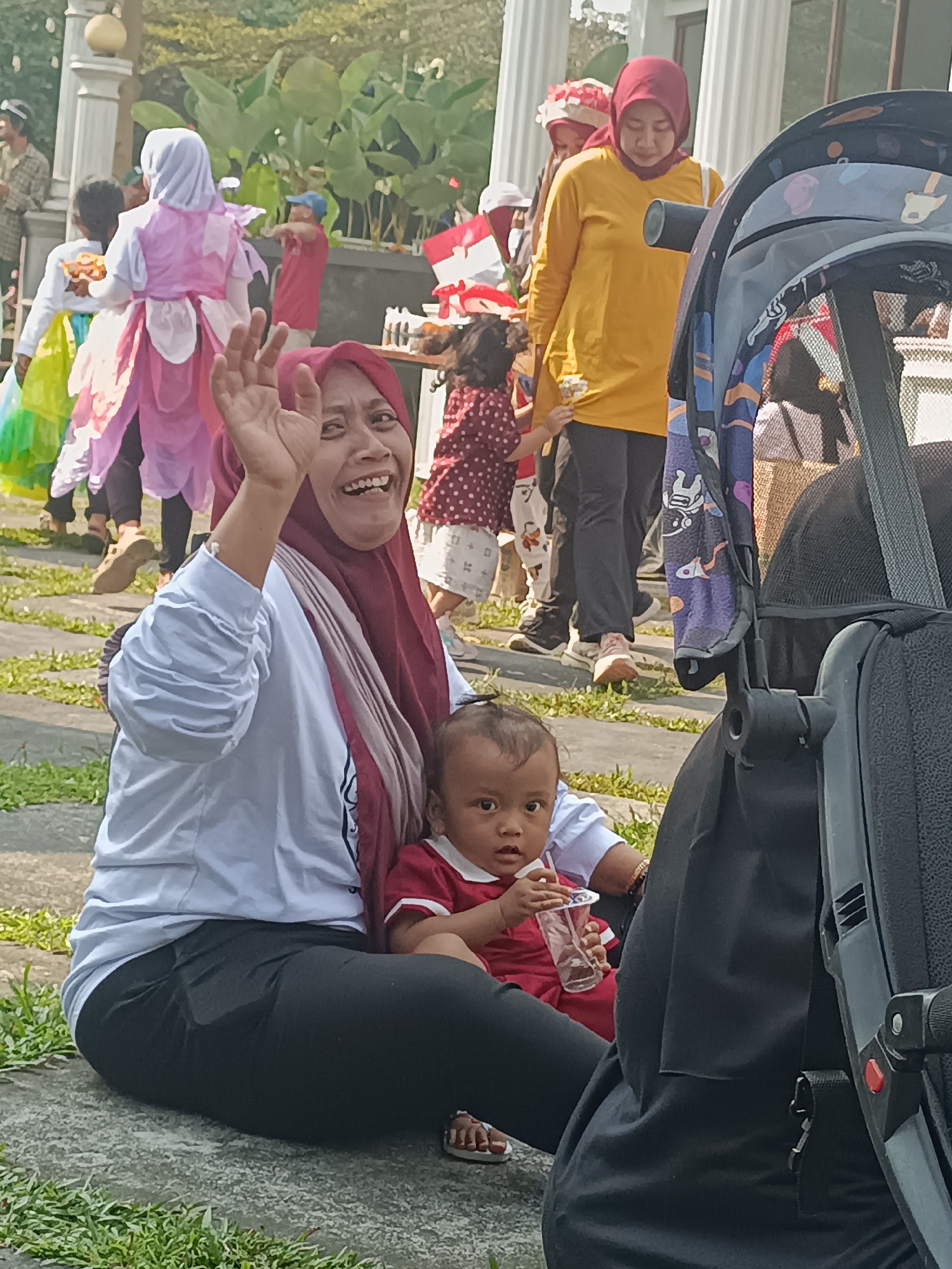 An outdoor scene at what appears to be a public event in Sleman, Indonesia, captures a smiling woman in a red hijab and white long-sleeved shirt waving her hand. She is seated on the ground next to a baby wearing a red shirt, who is holding a plastic cup with a straw. Behind them, several people are visible, including a woman in a yellow long-sleeved shirt and others in costumes. A stroller is partially visible in the foreground, on the right side of the image. The background shows lush green foliage and architectural elements, suggesting a park or a venue with landscaping. The ground is paved with stones and interspersed with grass. The overall atmosphere seems festive and casual.