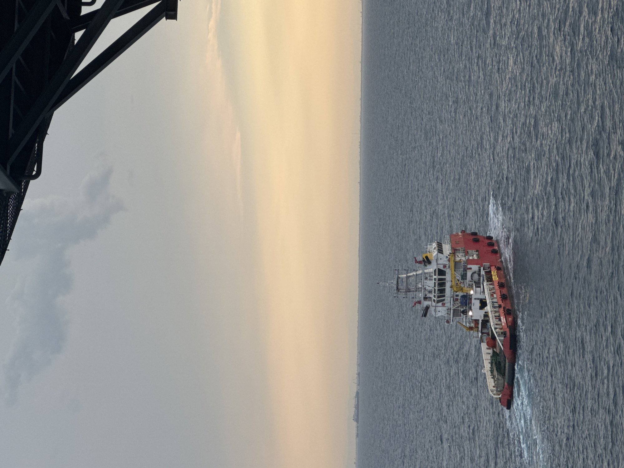 A high-angle, slightly tilted, medium shot captures a red and white offshore supply vessel traversing choppy grey water under an overcast sky. The vessel, positioned in the lower left quadrant of the frame, exhibits a clean white superstructure with red accents and a red hull. White foam trails indicate its movement. In the distance, along the horizon, a hazy, elongated landmass with faint structures and what appear to be industrial facilities or a port are visible. The sky transitions from a pale yellow near the horizon to a dull grey with scattered clouds. A dark, angular structure, possibly part of a larger bridge or platform, occupies the upper right corner of the image, partially obscuring the sky. The scene appears to be set in a maritime industrial area, though the specific city and country are not identifiable from the visual information.
