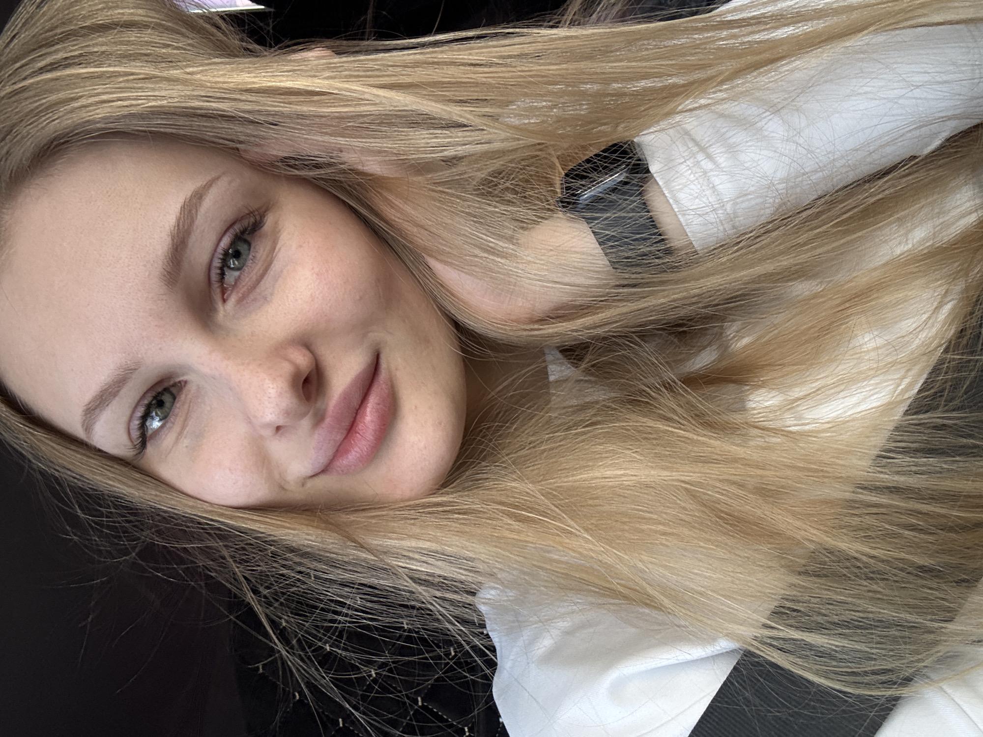 This close-up, tilted selfie features a young woman with long, straight blonde hair and light-colored eyes, appearing greenish-grey. She is looking directly at the camera with a subtle, pleasant smile, showing a calm and engaging expression. Her fair skin has visible freckles across her nose and cheeks. Her hair cascades over her left arm and shoulder, partially obscuring a black smartwatch or fitness tracker on her wrist. She is wearing a white top or shirt, with a dark strap or element visible beneath her hair and clothing in the lower right. The background is dark and out of focus, providing no specific details about the scene or setting. The image appears to be a casual, personal portrait, likely taken indoors. No text is visible, and the time of day or weather cannot be determined from the image.