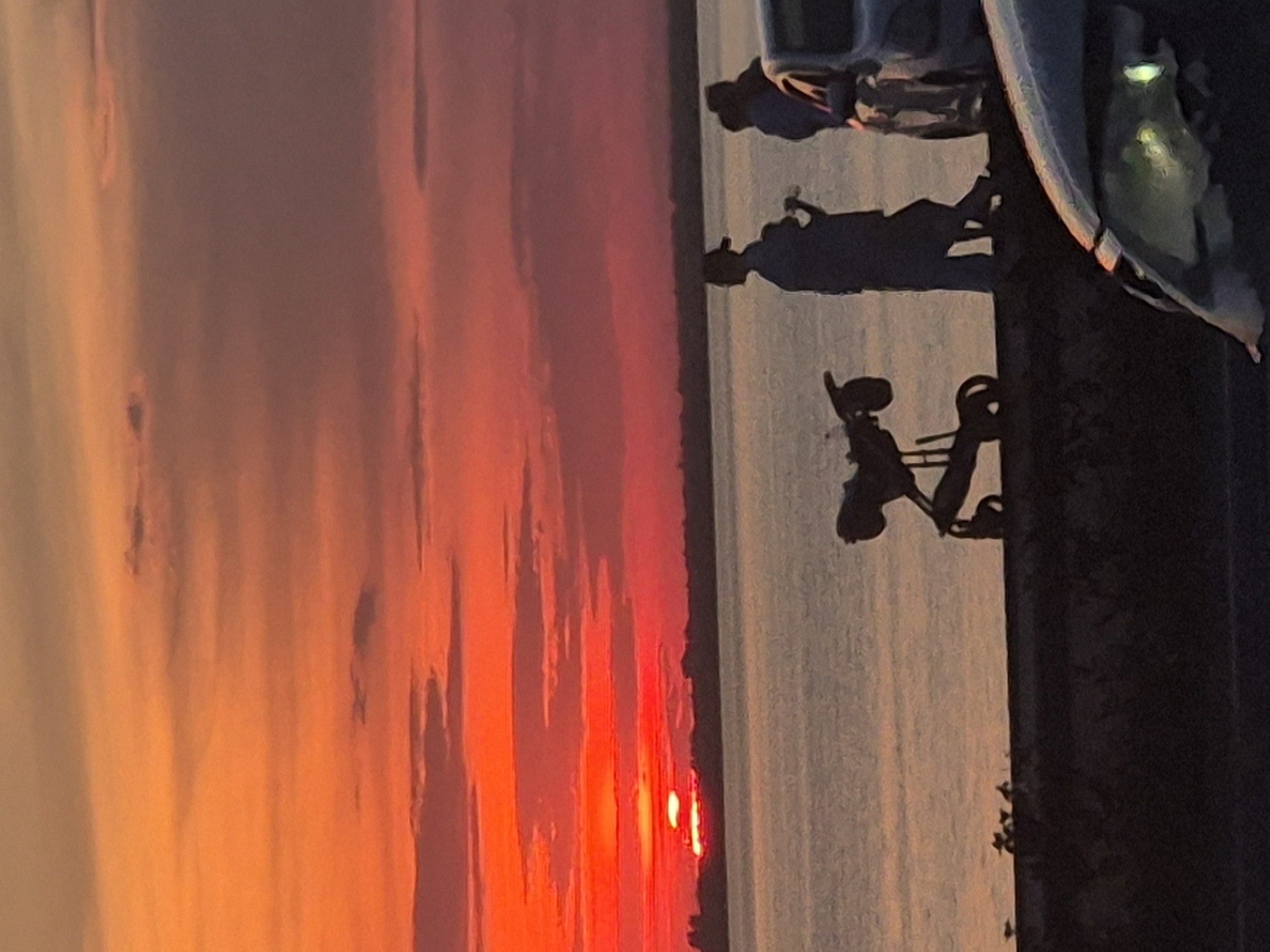 The image depicts a sunset over a body of water. Silhouetted figures stand on a walkway along the water's edge. To the left, a stroller-like object is visible. In the background, a distant shoreline is outlined against the vibrant orange and red hues of the sky. The water is calm and reflects the colors of the sunset. The scene appears to be a public area, possibly a promenade or waterfront park.